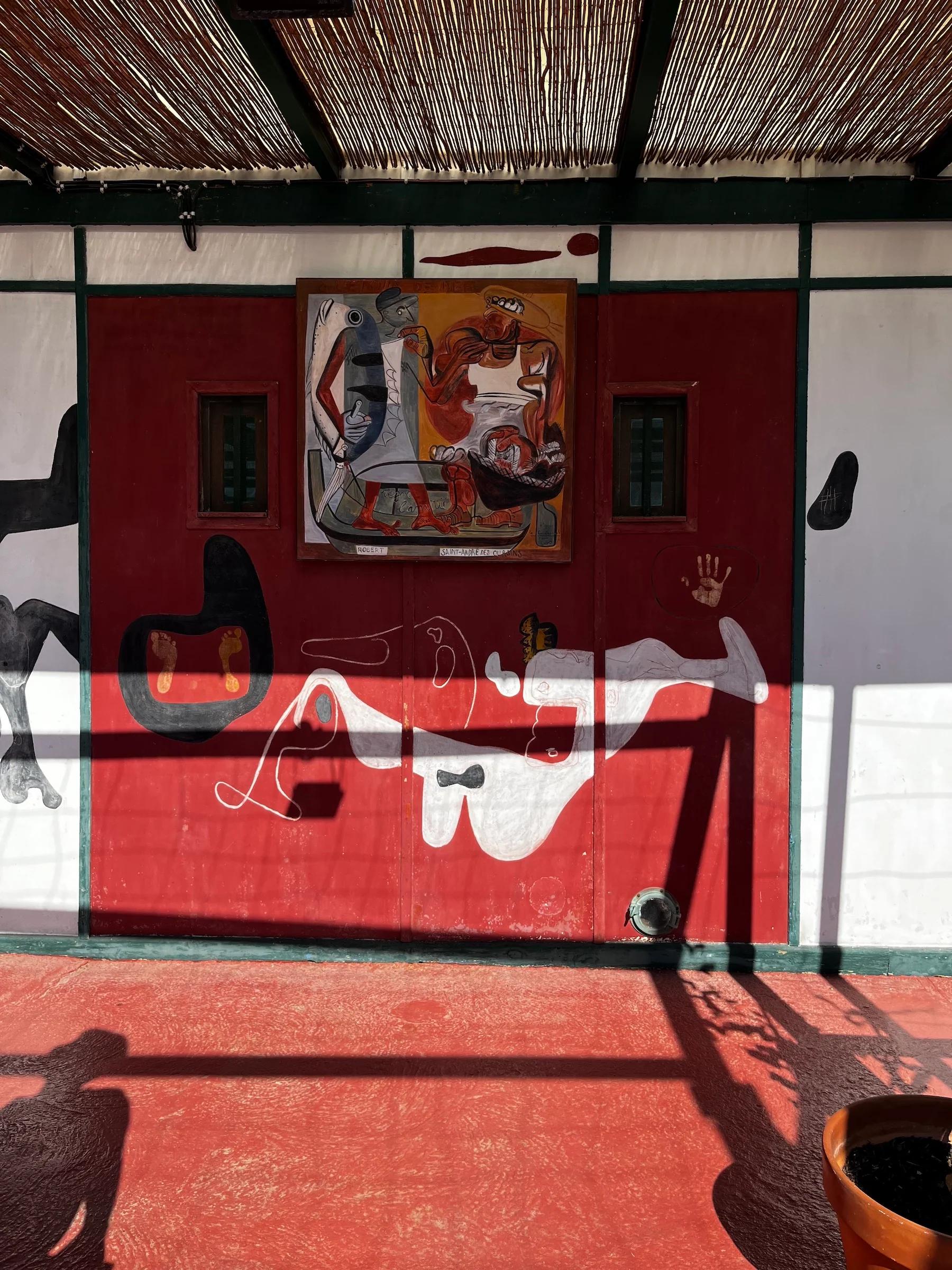 Pergola mural in Marseille with abstract art featuring human and animal forms in white, red, and black. Central framed painting flanked by small windows.