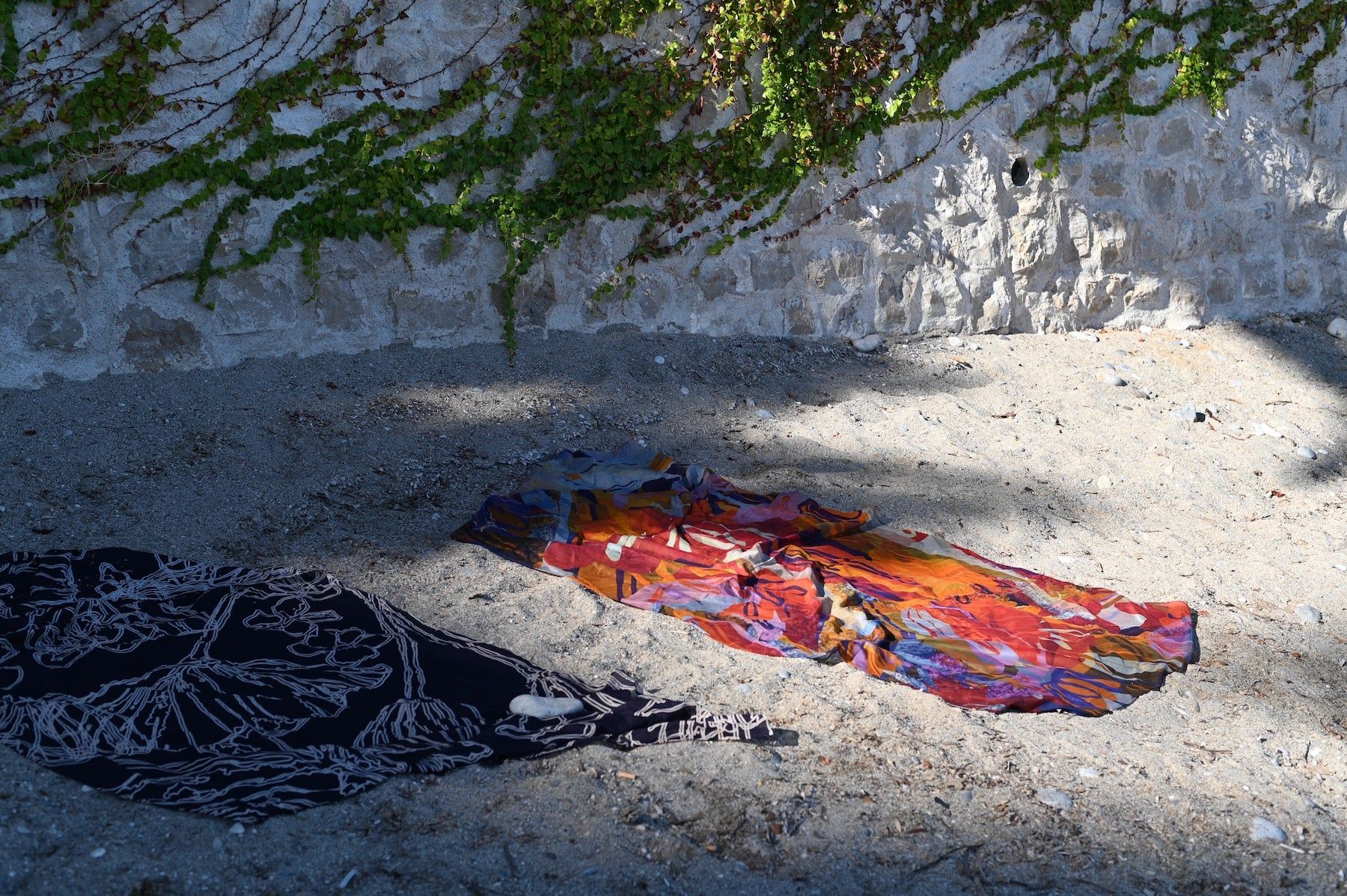 Two colourful beach towels on sandy shore. One multicoloured, one black and white. Green vines hanging from stone wall casting shadows.