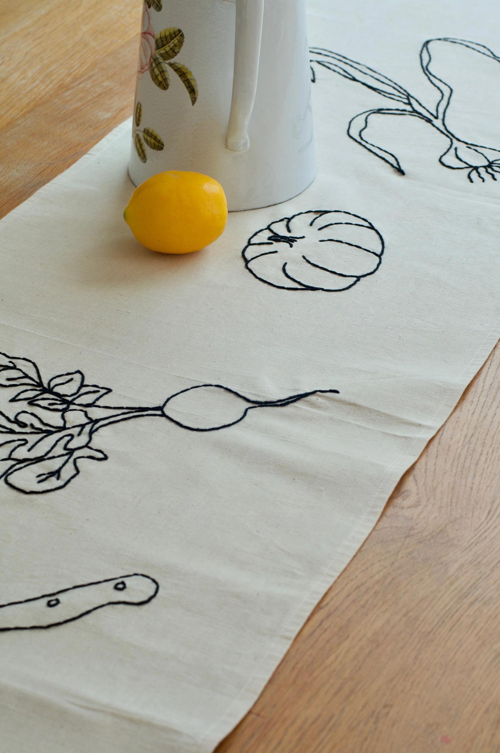 White table runner with embroidered vegetable outlines on wooden table. White jug with floral design and lemon on runner.
