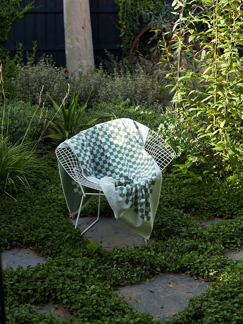 A green and white checkered cotton blanket draped over a white metal chair in a lush garden, surrounded by plants. Sunlight highlights the peaceful setting.