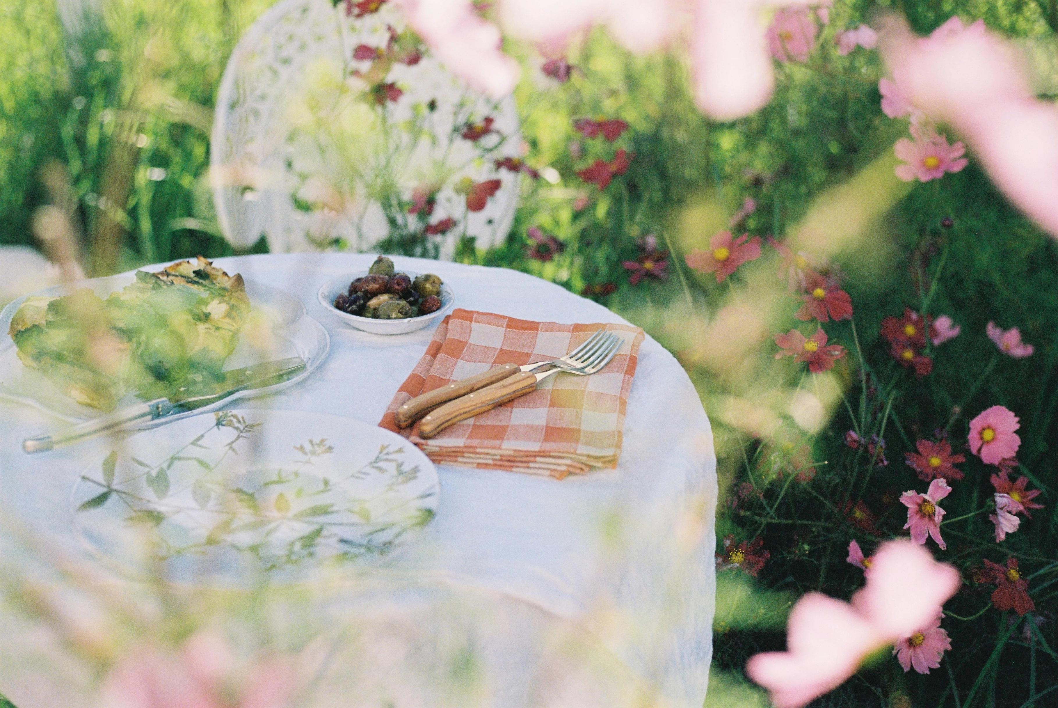 Outdoor table with white cloth and floral plates. Orange-checked napkin holds wooden utensils. Olives, salad, and green frittata nearby. Pink flowers surround the scene.