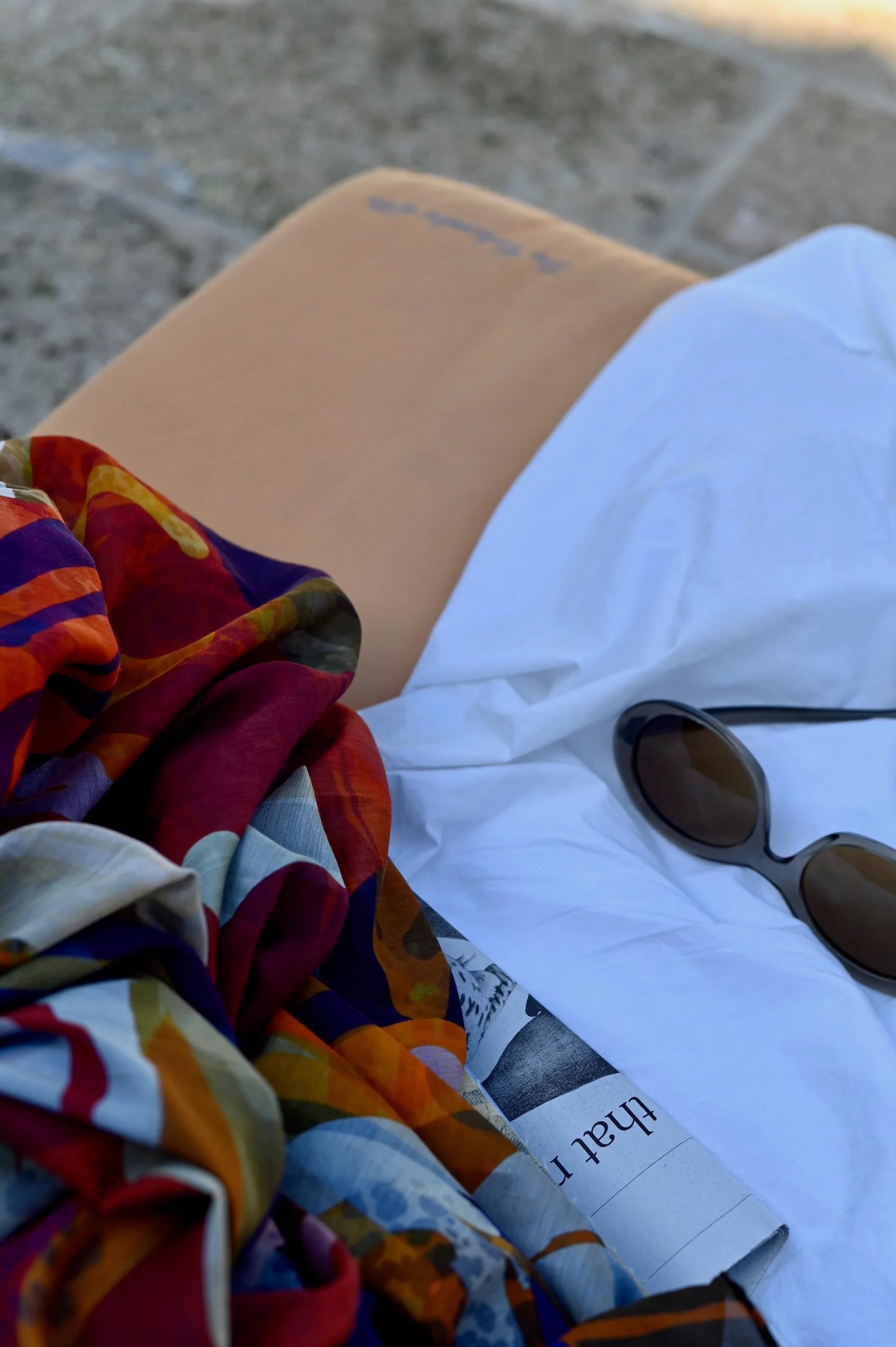 Close-up of tan cushion with newspaper, sunglasses, and colourful fabric on white shirt. Woven bags nearby.