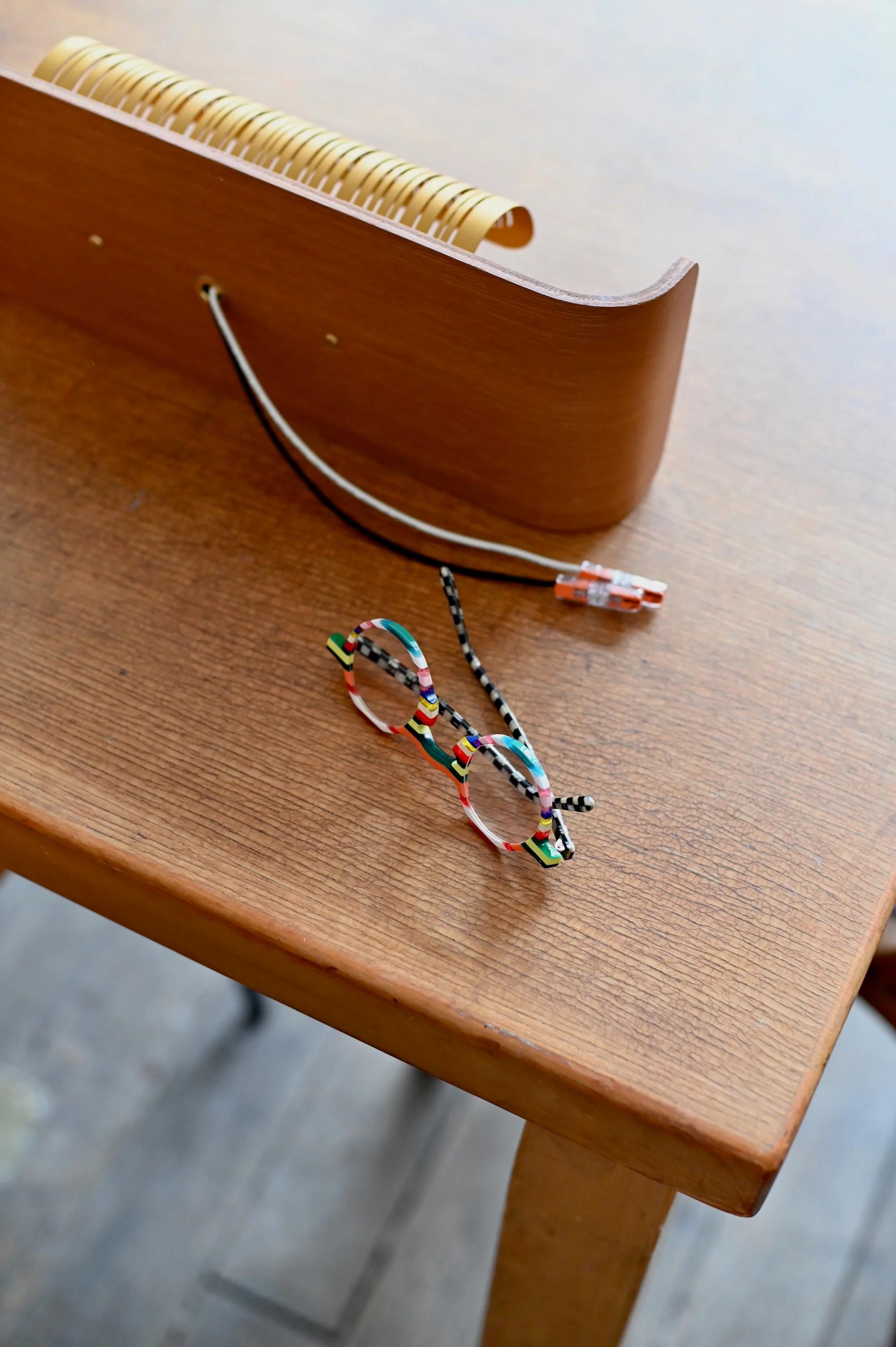 Colourful glasses with beaded chain on table. Wooden desk organiser and black-white cord nearby. Blurred background.