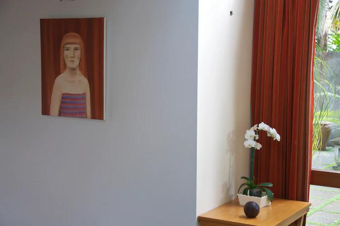 Minimalist room with red-haired portrait. Small table displays Rachel Carley's white orchid pot and dark sphere. Red curtains frame large window.