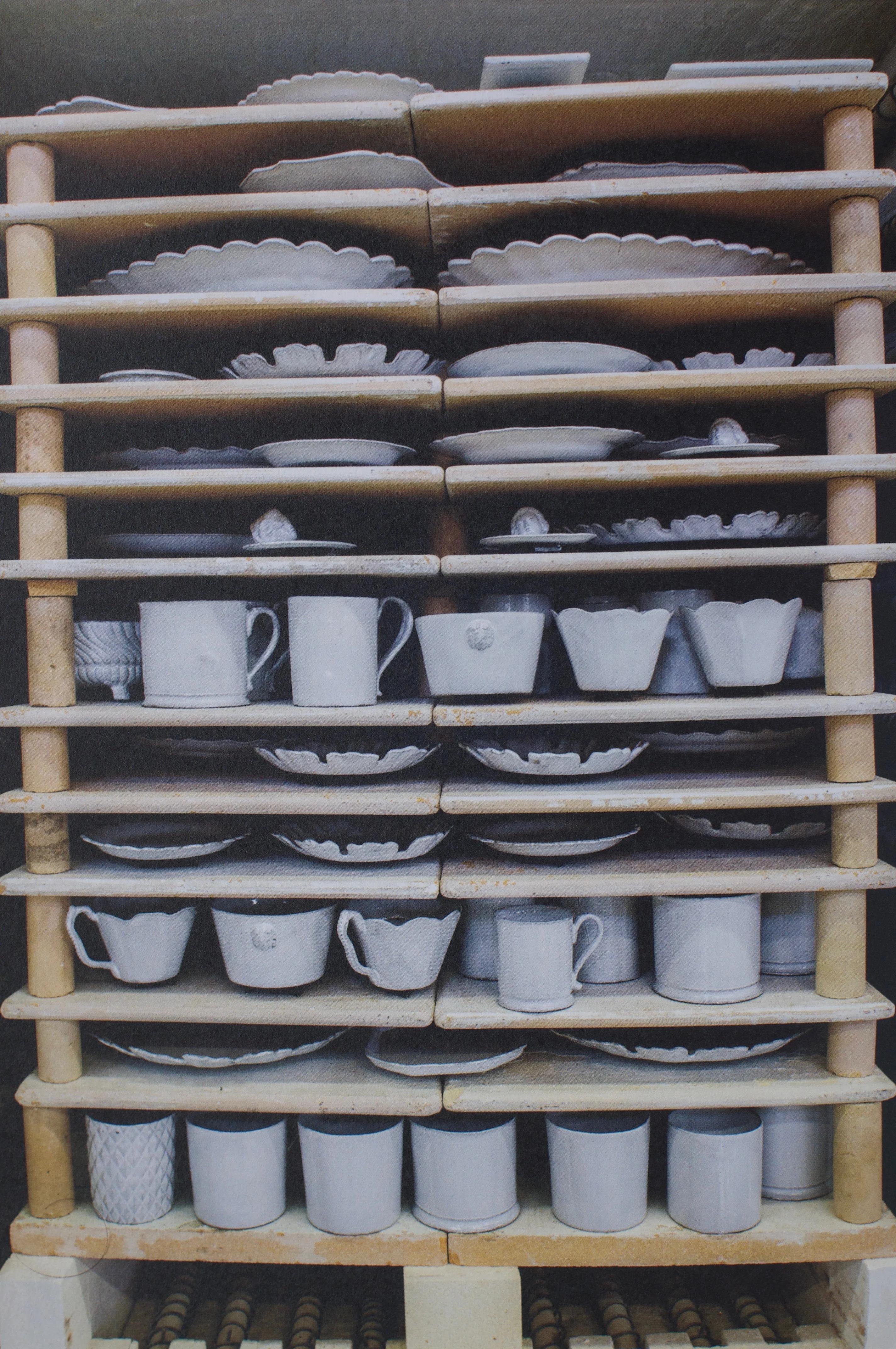 A wooden shelf displays white ceramic dishes, cups, and bowls. The handcrafted pottery is neatly arranged in various shapes and sizes.