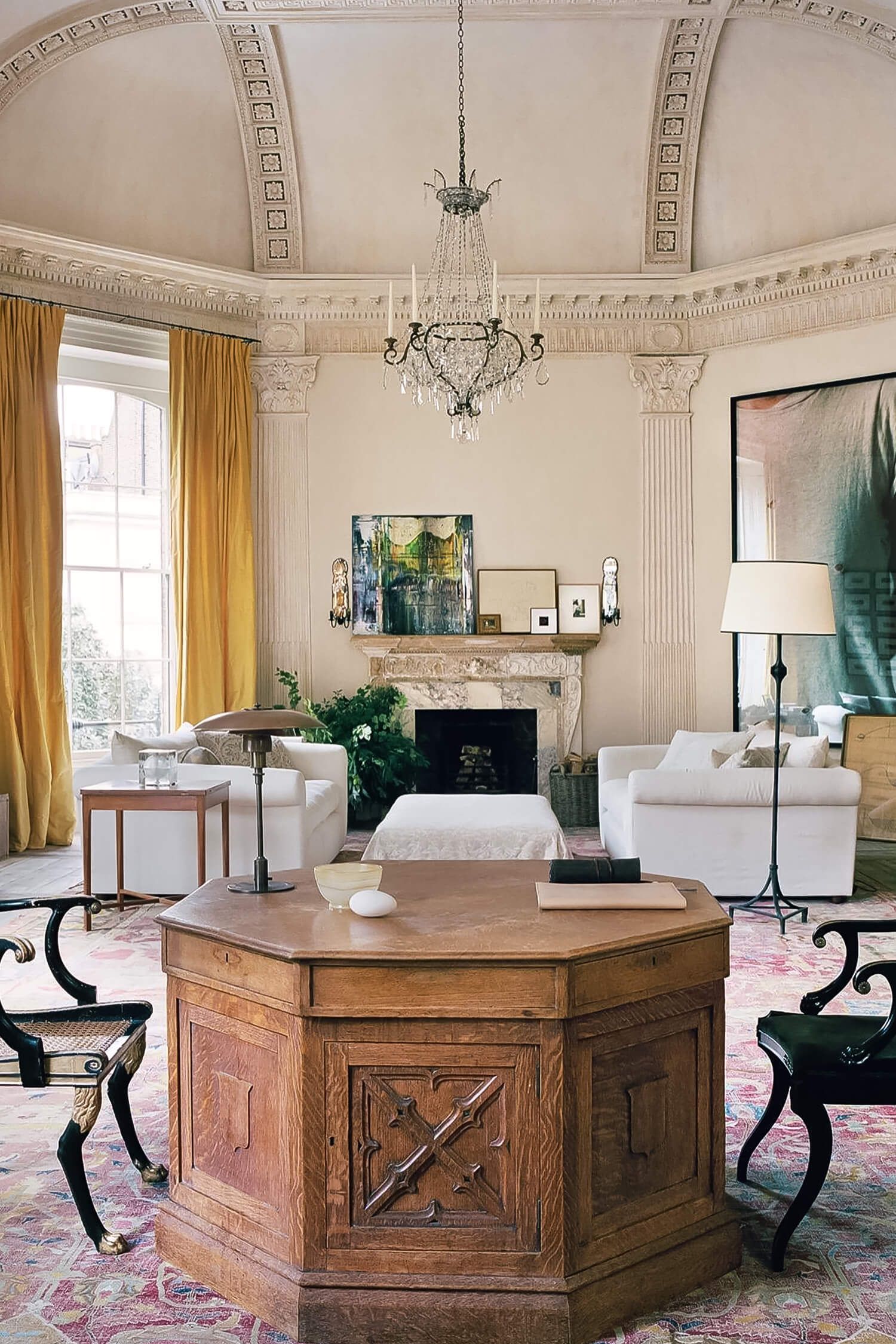 Grand living room with ornate ceiling. Large windows, chandelier, fireplace with art. Mix of classic and modern furniture.