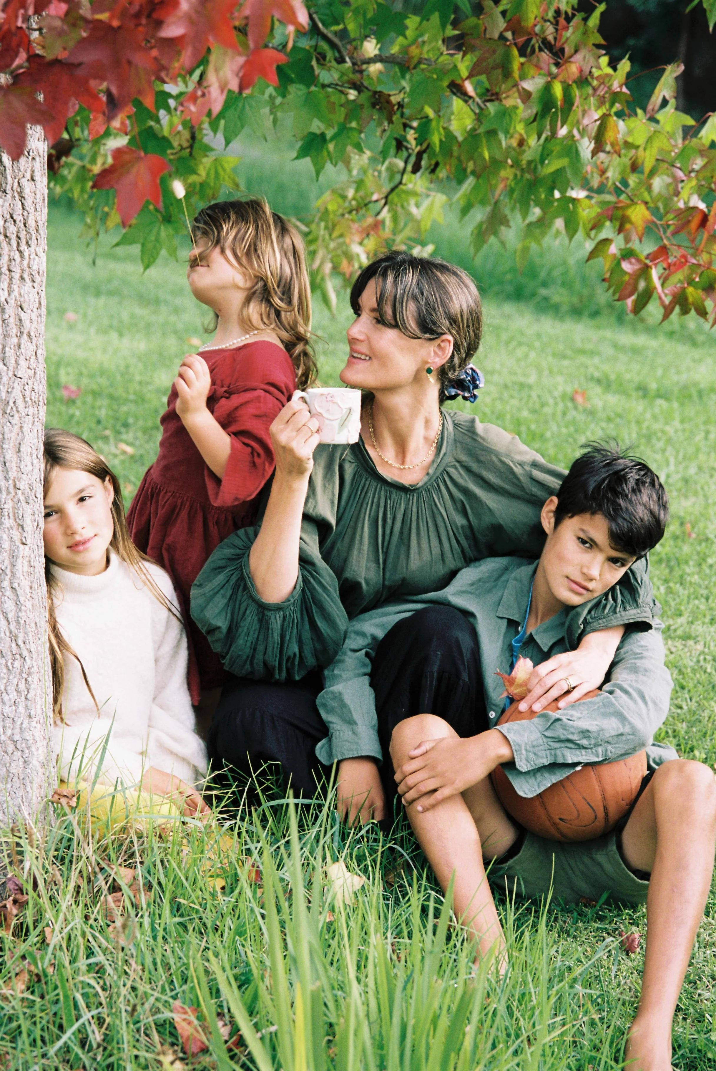 A woman sits on grass under a tree with three children. One girl in red leans on the tree, a boy in green holds a basketball, and a girl in white sits nearby. The woman smiles, holding a mug.