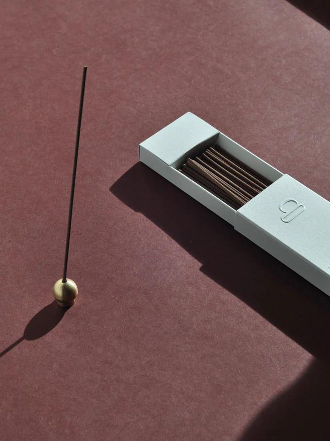 Lit incense in brass holder on maroon surface. Open white box with unused incense sticks nearby.