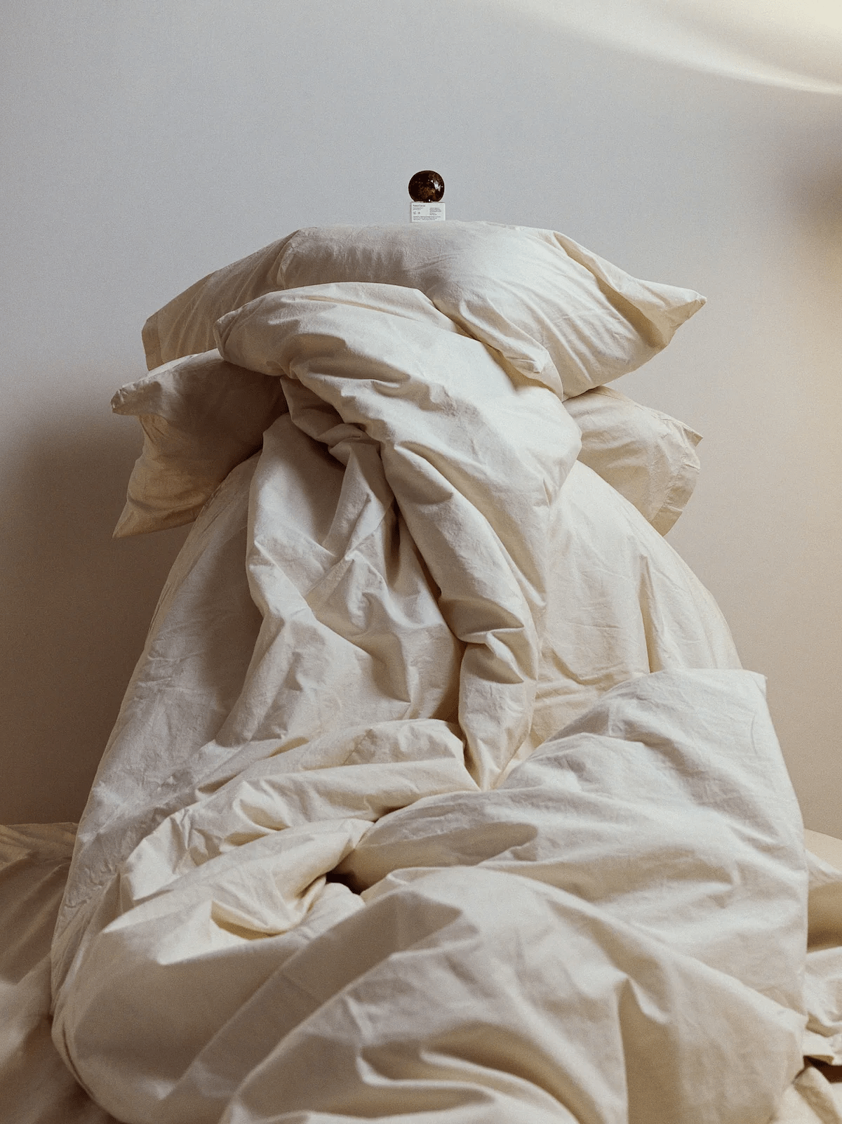 Disheveled white bed linens with small spherical object on white card at peak. Light-coloured wall background.