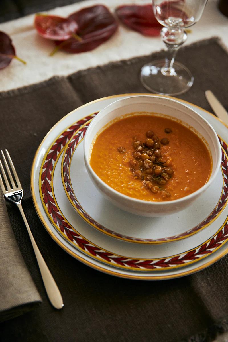 A bowl of pumpkin soup with lentils on decorative plates. Fork and knife rest on a brown napkin. Wine glass and autumn leaves complete the elegant setting.