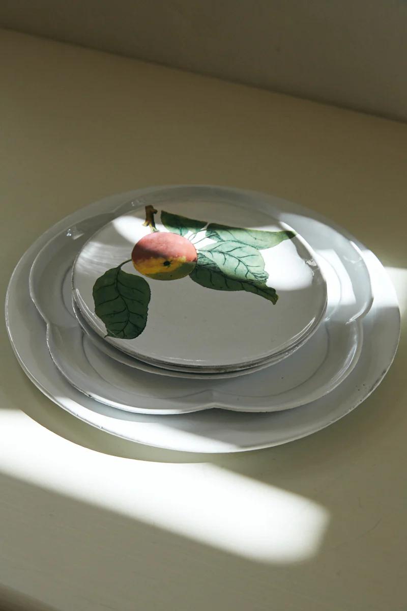 Stack of white plates, top plate with colourful fruit design. Soft natural light on light surface.