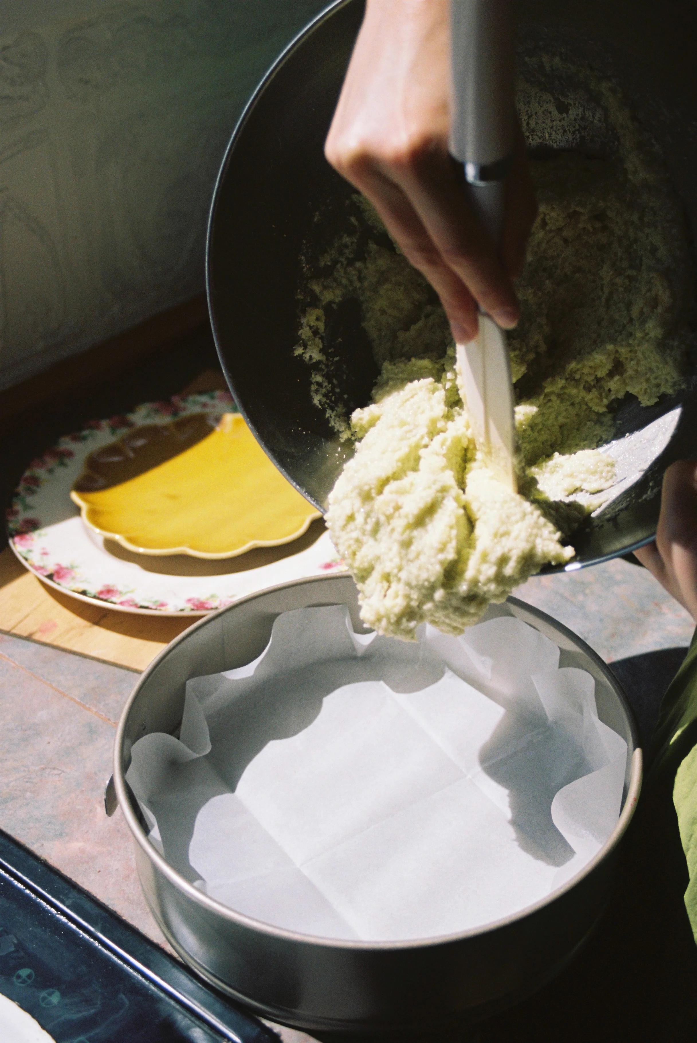 Person transferring batter to lined cake pan. Coconut Raspberry cake kit and yellow floral plate in background.