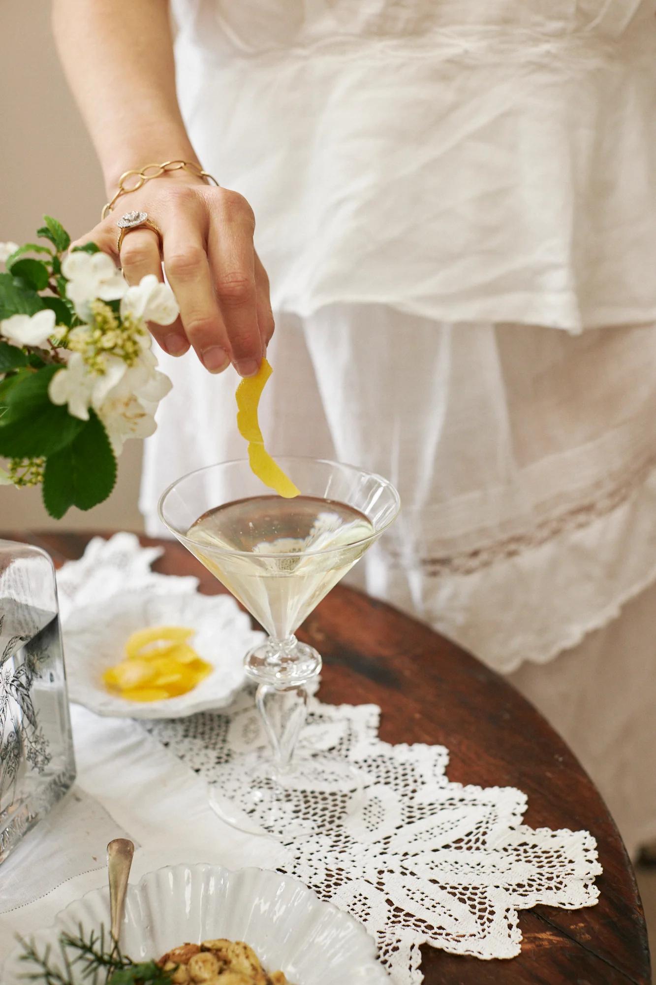 A hand adds a lemon twist to a martini on a lace-covered table. White flowers and garnish dishes are nearby, creating an elegant cocktail setting.