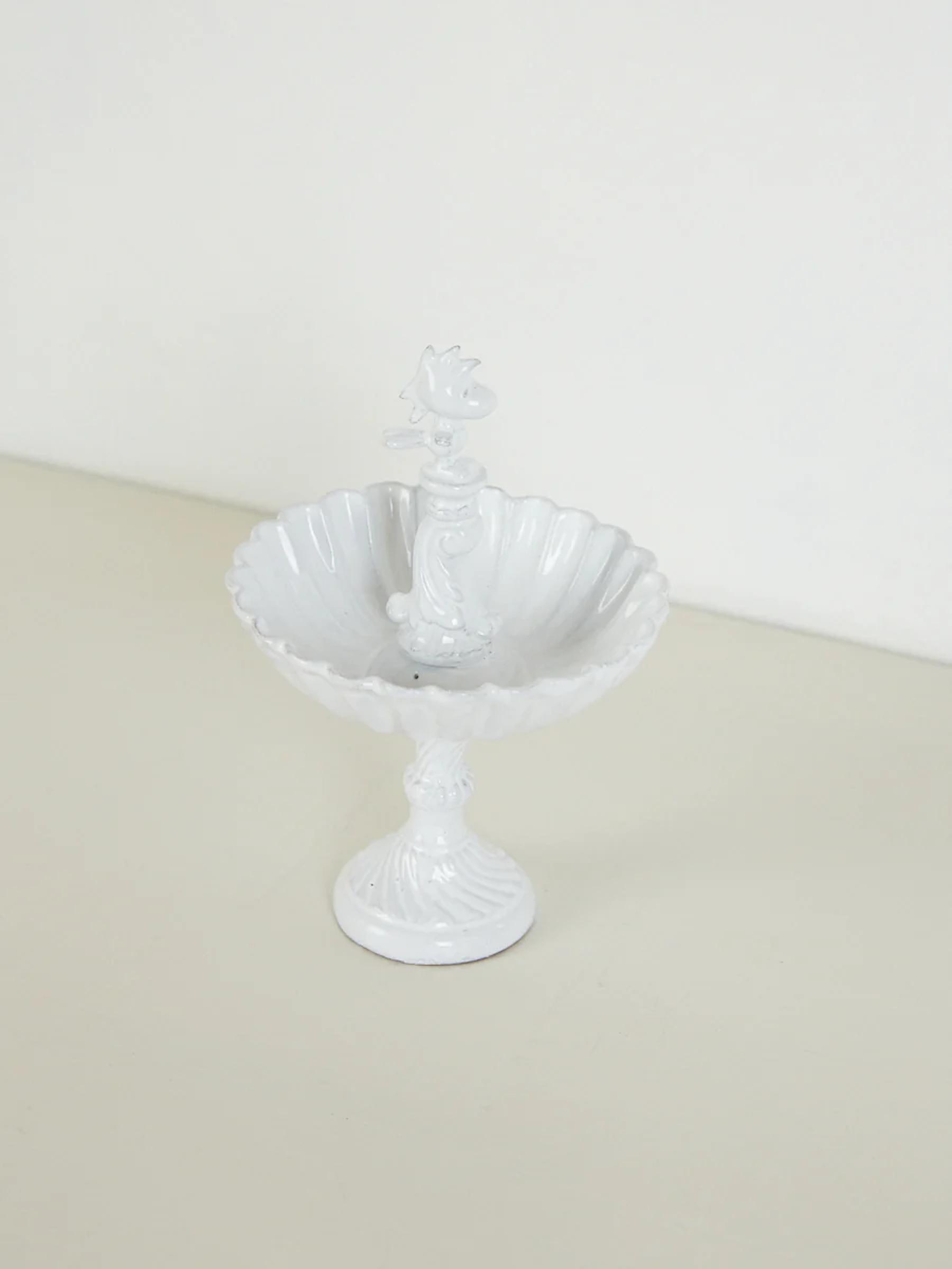 White ceramic bowl with scalloped edge, ornate stem, bird on top. Pedestal base. Light, plain background.