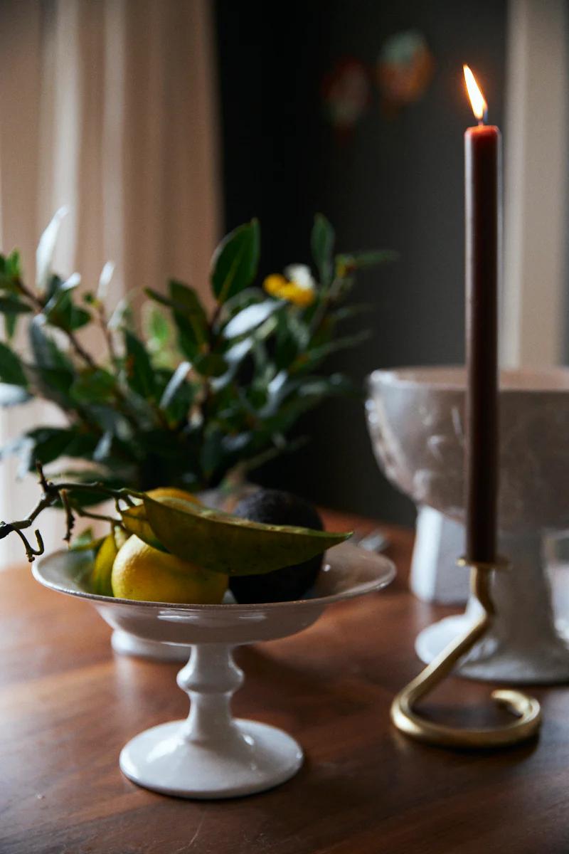 Lit candle by fruit bowl on wooden table. Cheese platter nearby. Cosy, dim atmosphere.