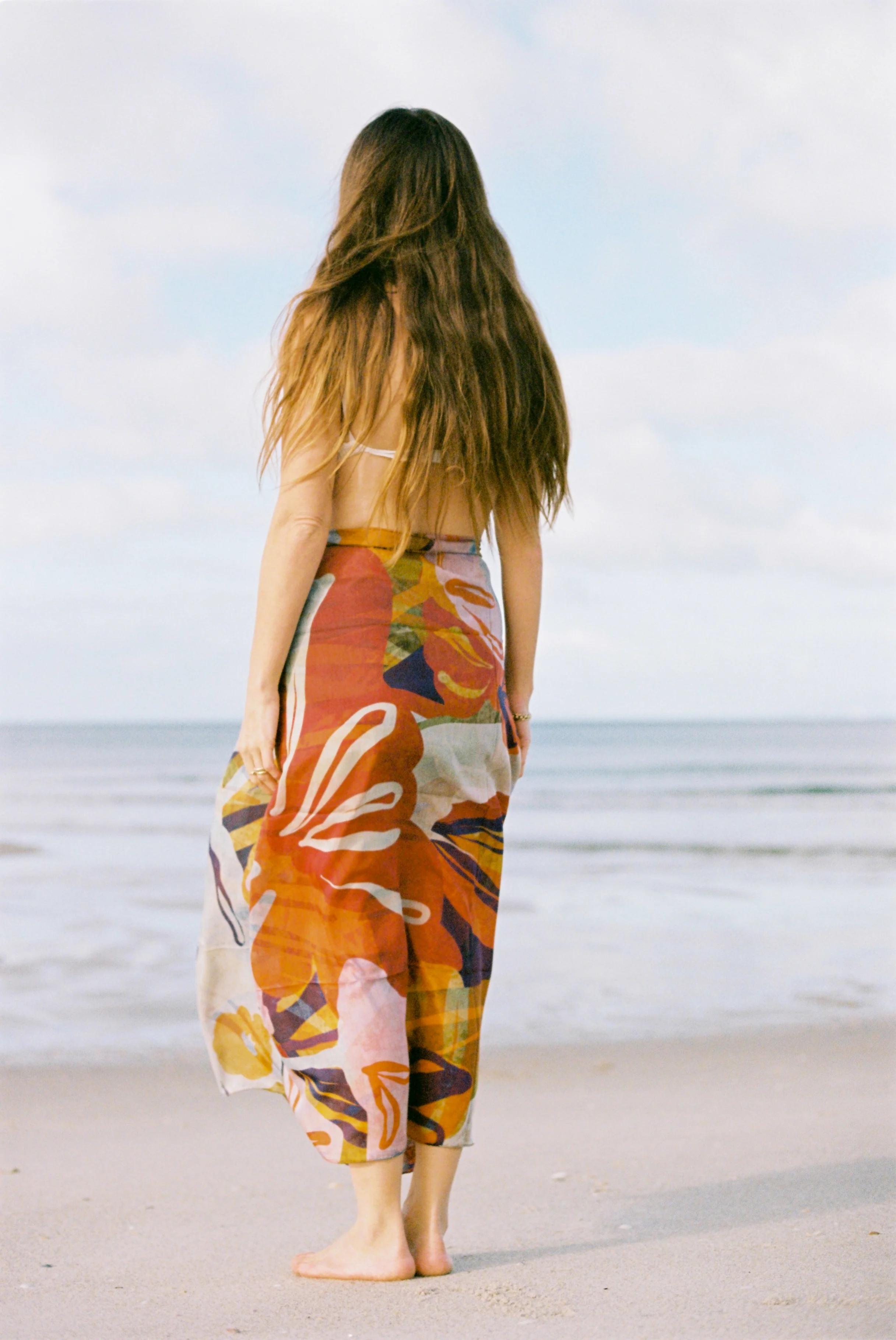 Person with long brown hair on sandy beach facing ocean. Wearing colourful, patterned flowing dress. Partly cloudy sky, calm water.