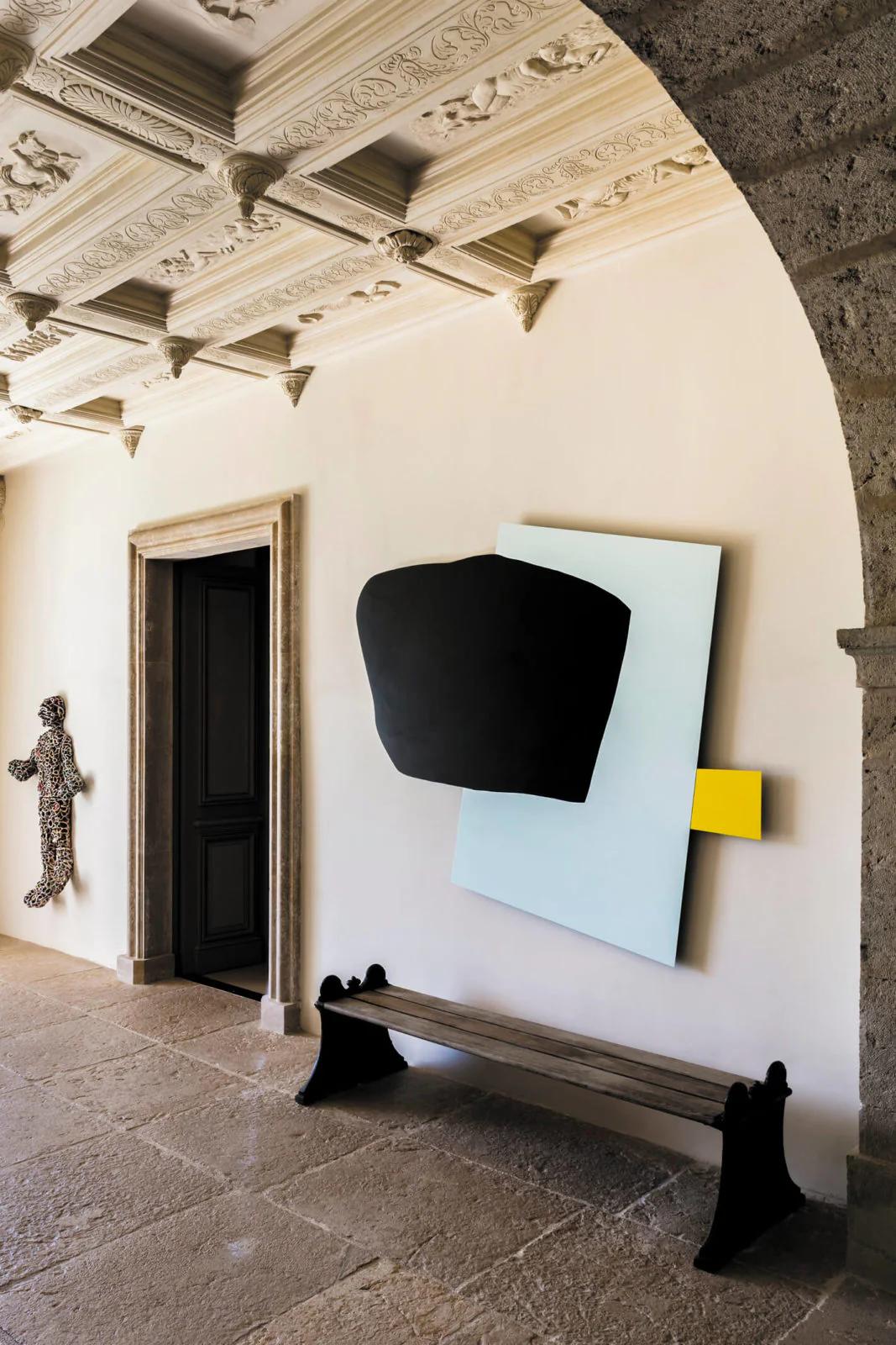 A stone-floored hallway with ornate ceiling and black door. Abstract art hangs above a rustic bench, evoking south of France charm.