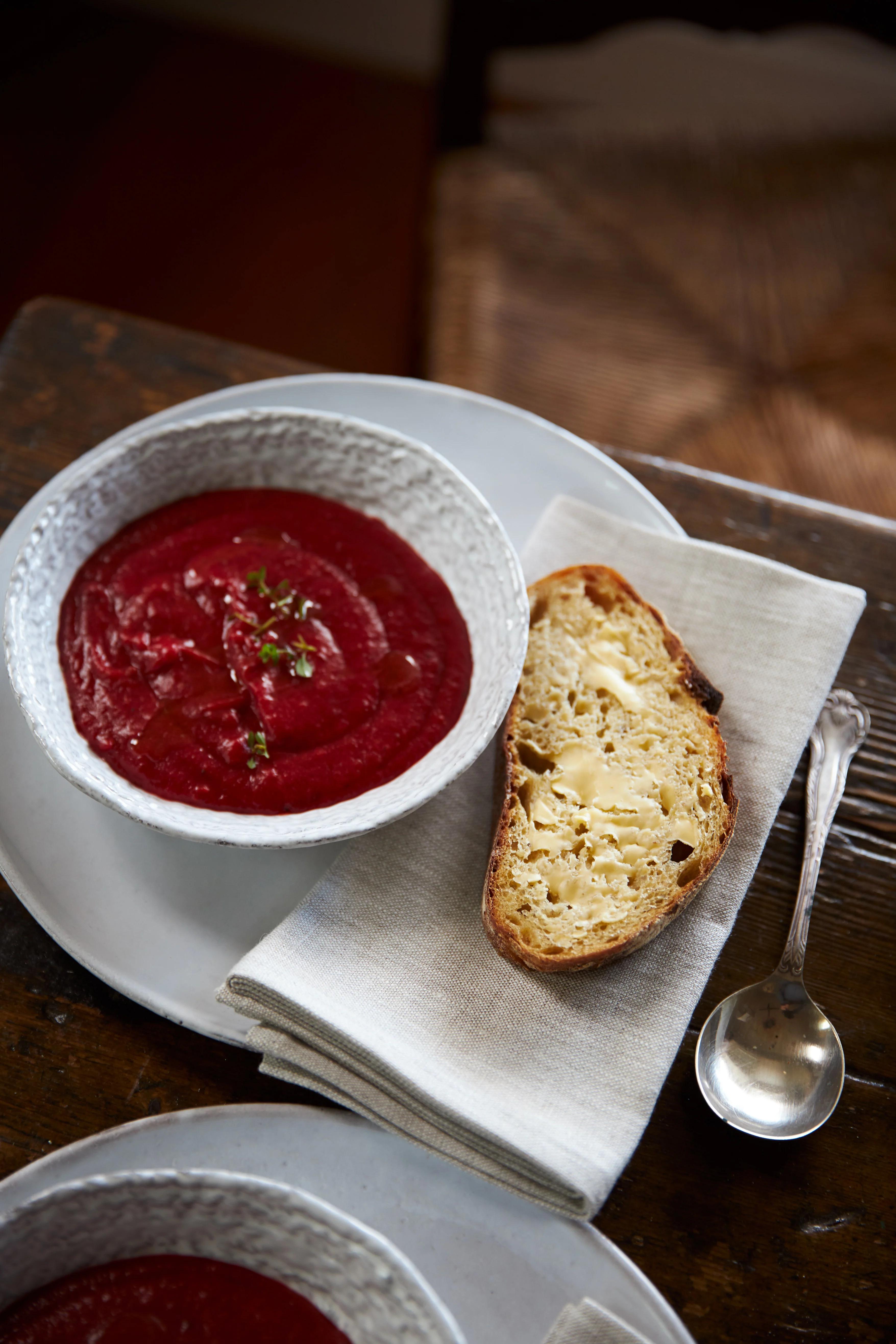 A bowl of creamy beetroot soup garnished with herbs sits on a white napkin, with a slice of buttered bread and a spoon beside it. The setup on a wooden table feels perfect for a rejuvenating meal.