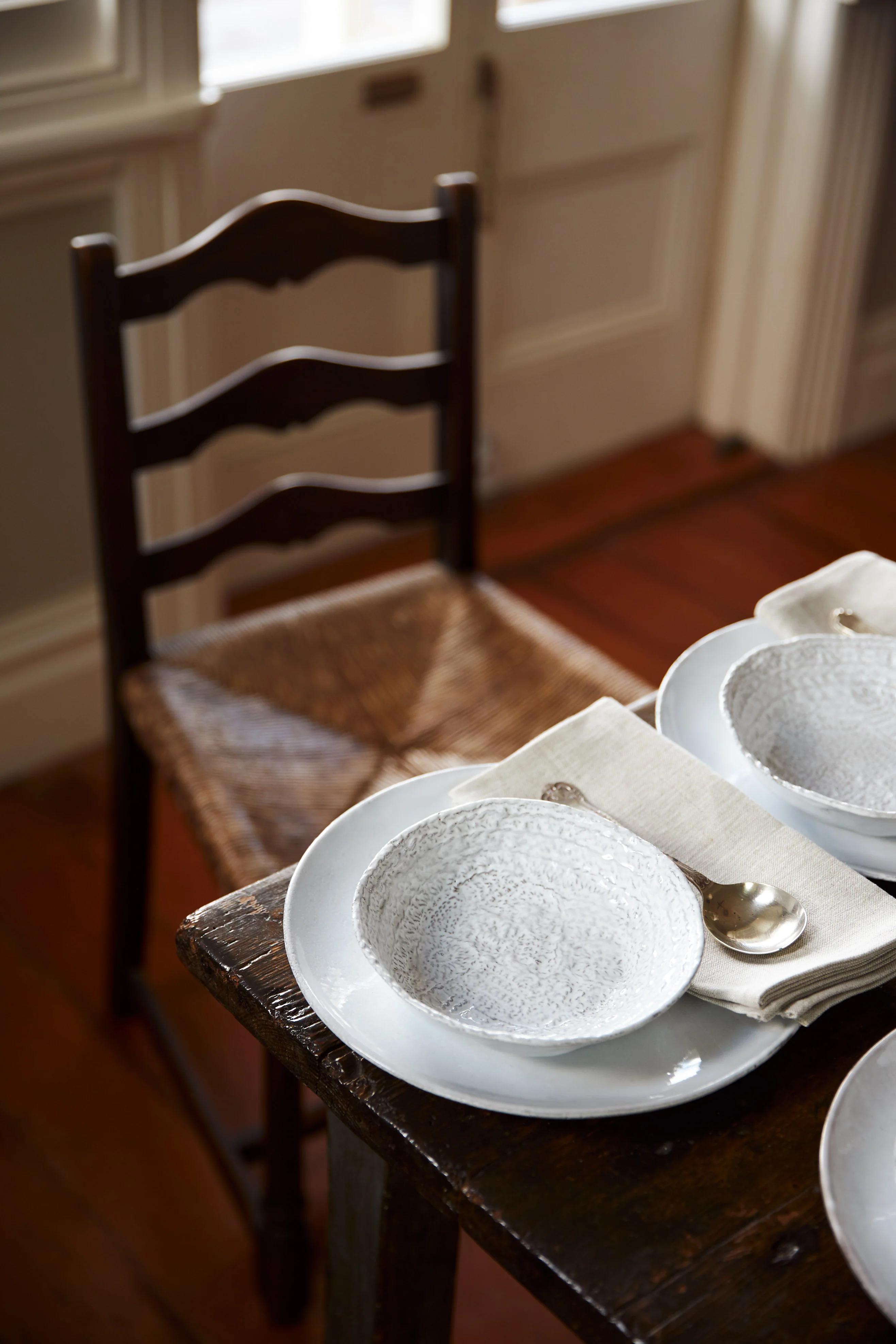 A rustic wooden dining table with white dishware, cloth napkins, and spoons, next to a woven wooden chair. Sunlight creates a warm, inviting setting.