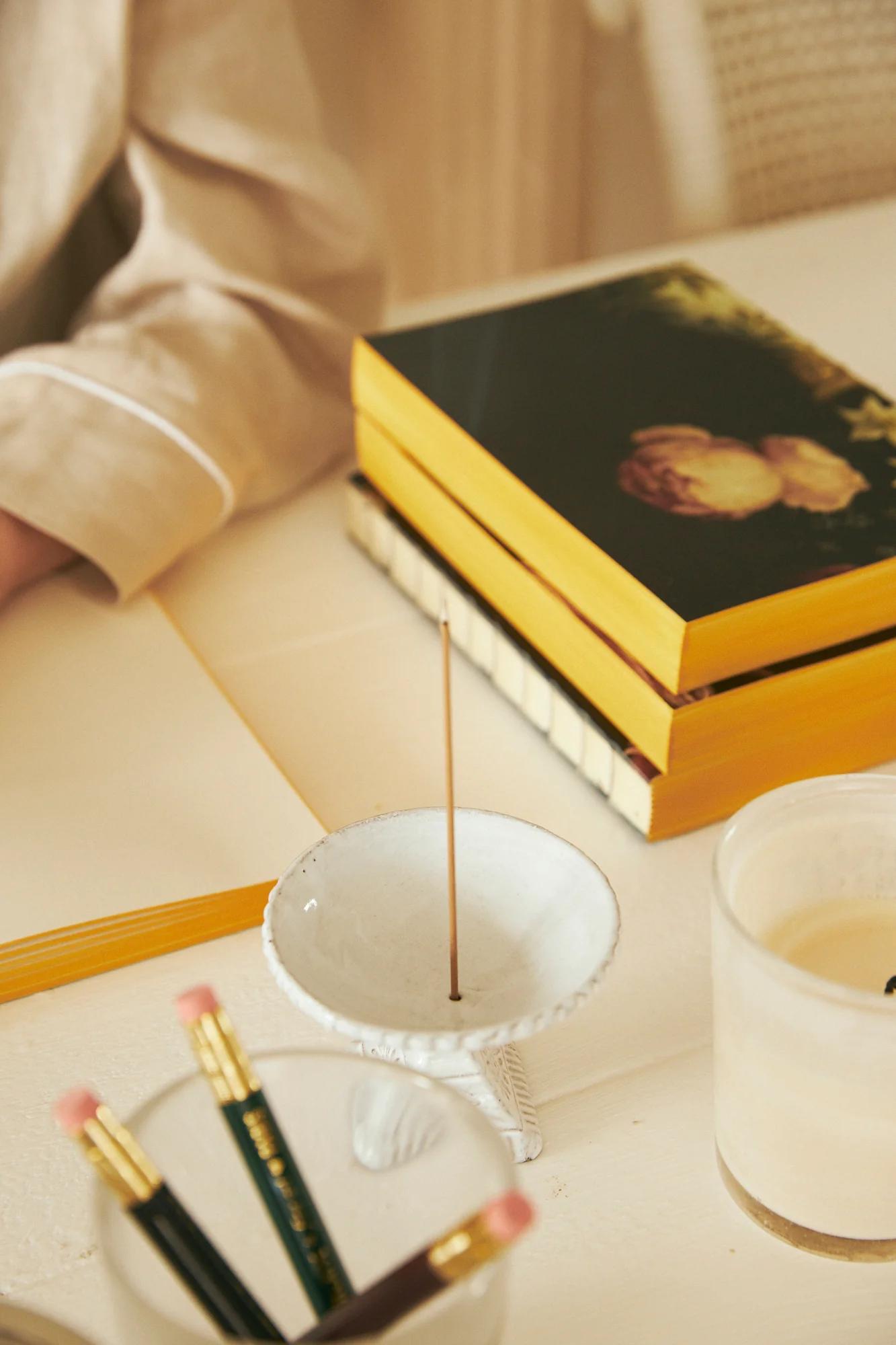 Desk with yellow books, person writing cards, burning incense, pencil container, and lit candle.