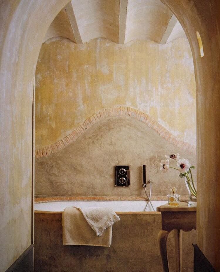 Rustic bathroom with stone tub and textured walls. Curved archway entrance. Wooden chair holds flowers. Astier de Villatte ceramic soap dishes add charm.