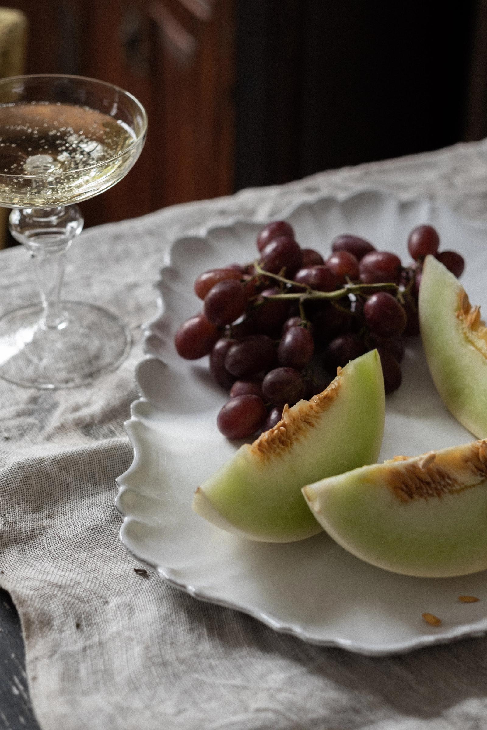 a ceramic platter with grapes and melon sits on a table next to a champagne glass