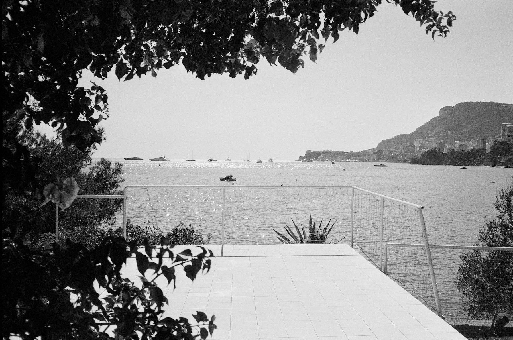 Black and white coastal scene with tiled terrace overlooking calm waters and distant yachts. Framed by foliage with visible mountainous coastline.