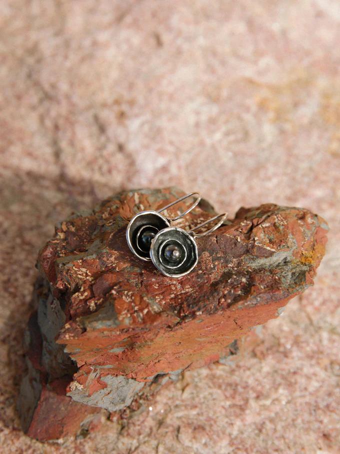 Silver earrings with dark centres on textured red-brown rock. Background highlights natural formations, emphasising organic-inspired jewellery design.