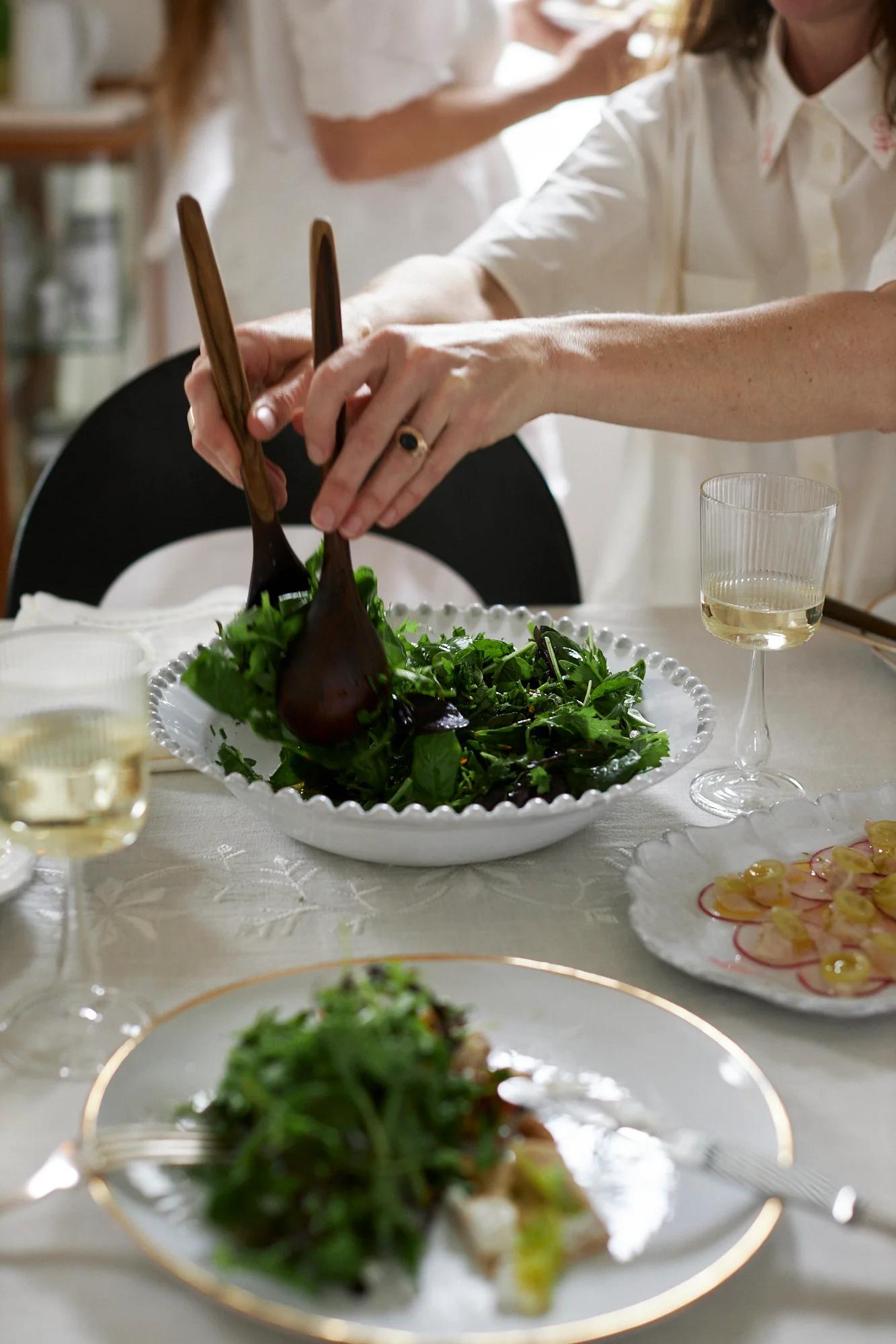 Someone serves green salad from a large bowl. The bright room shows plates of food, wine glasses, and another person in the background.