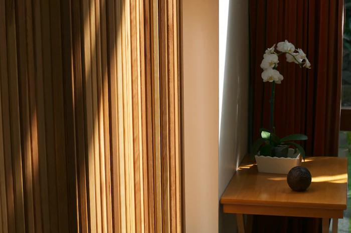 Serene corner with wooden slats and sunlit table. White vase holds blooming orchids, resembling a ceramic artist's peaceful retreat.