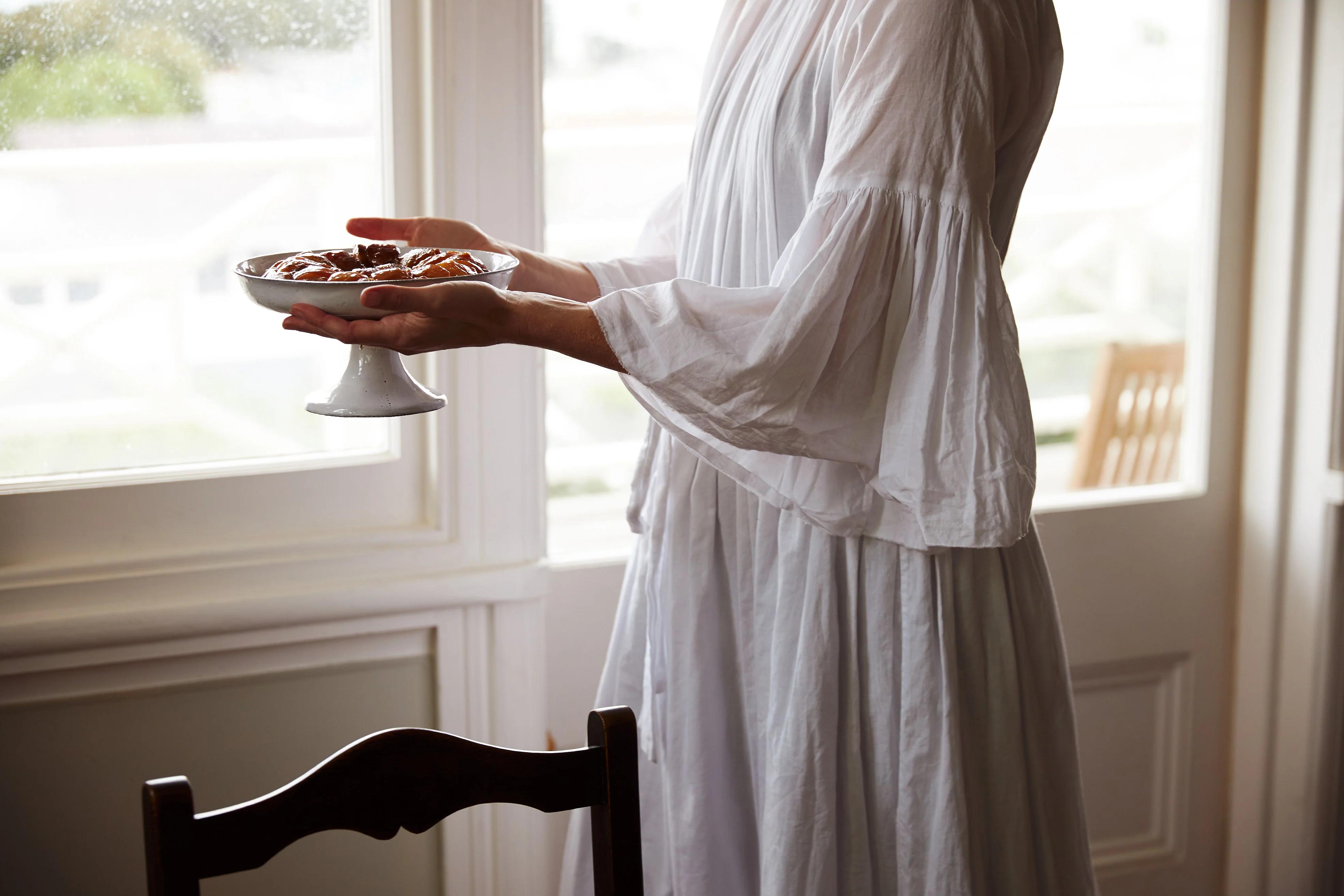 Person in flowing white dress holds pedestal tray of icing sugar-dusted baked goods near window. Softly lit room with wooden chair in foreground.