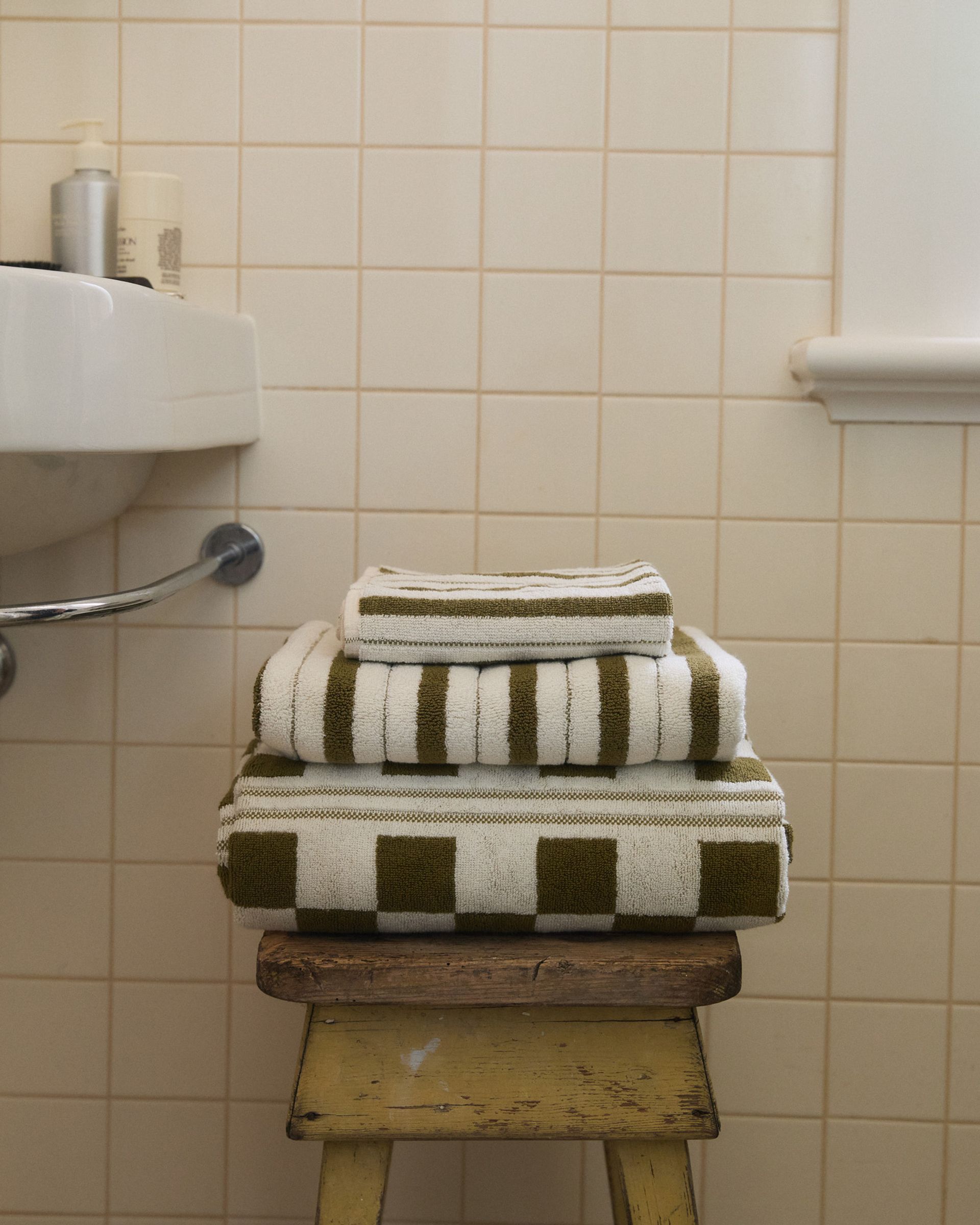three towels sit folded on top of a wooden vintage stool in a white tiled bathroom