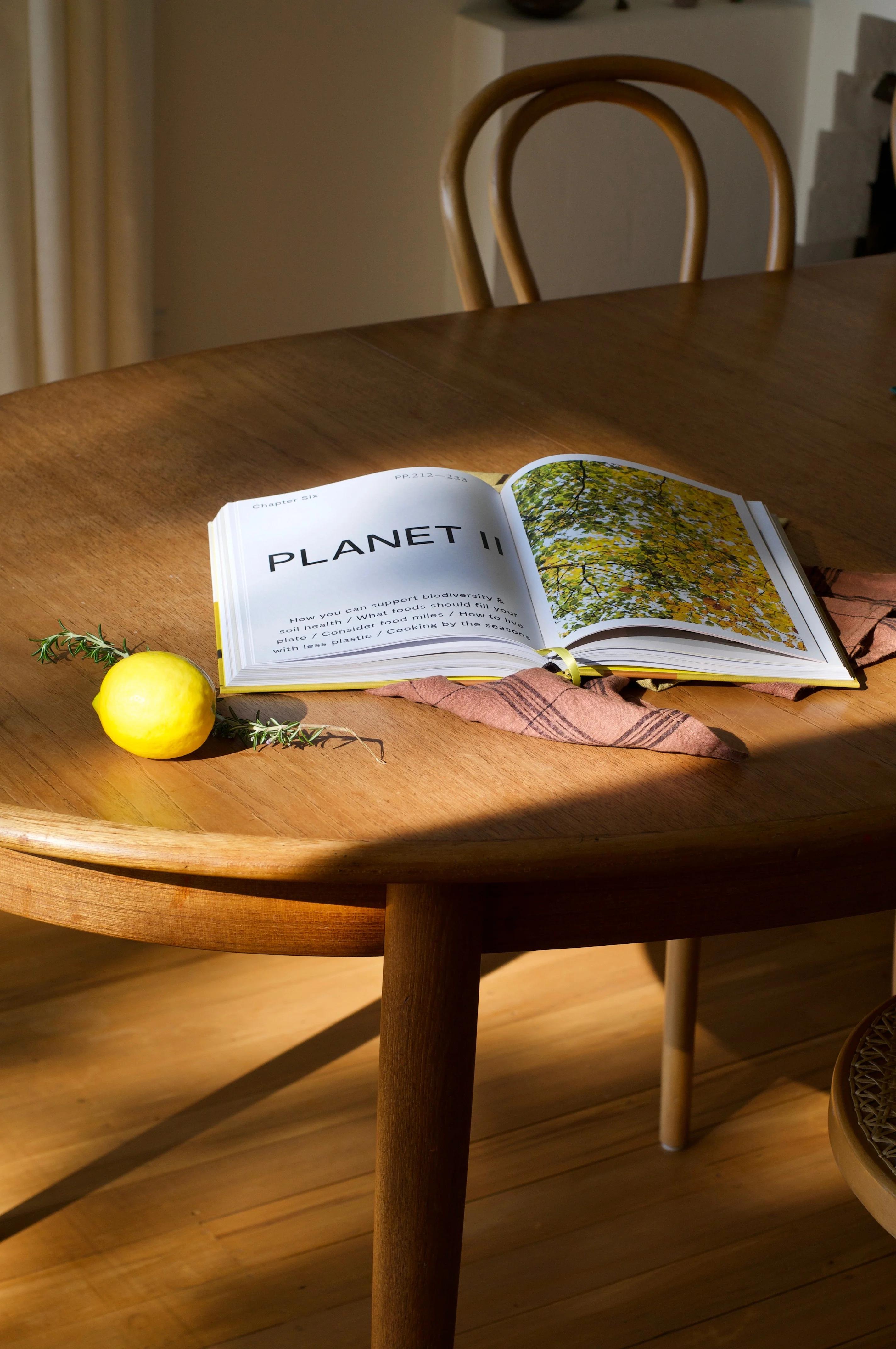 Open "Planet II" book on wooden table showing floral images. Lemon, greens, folded cloth nearby. Sunlit scene.