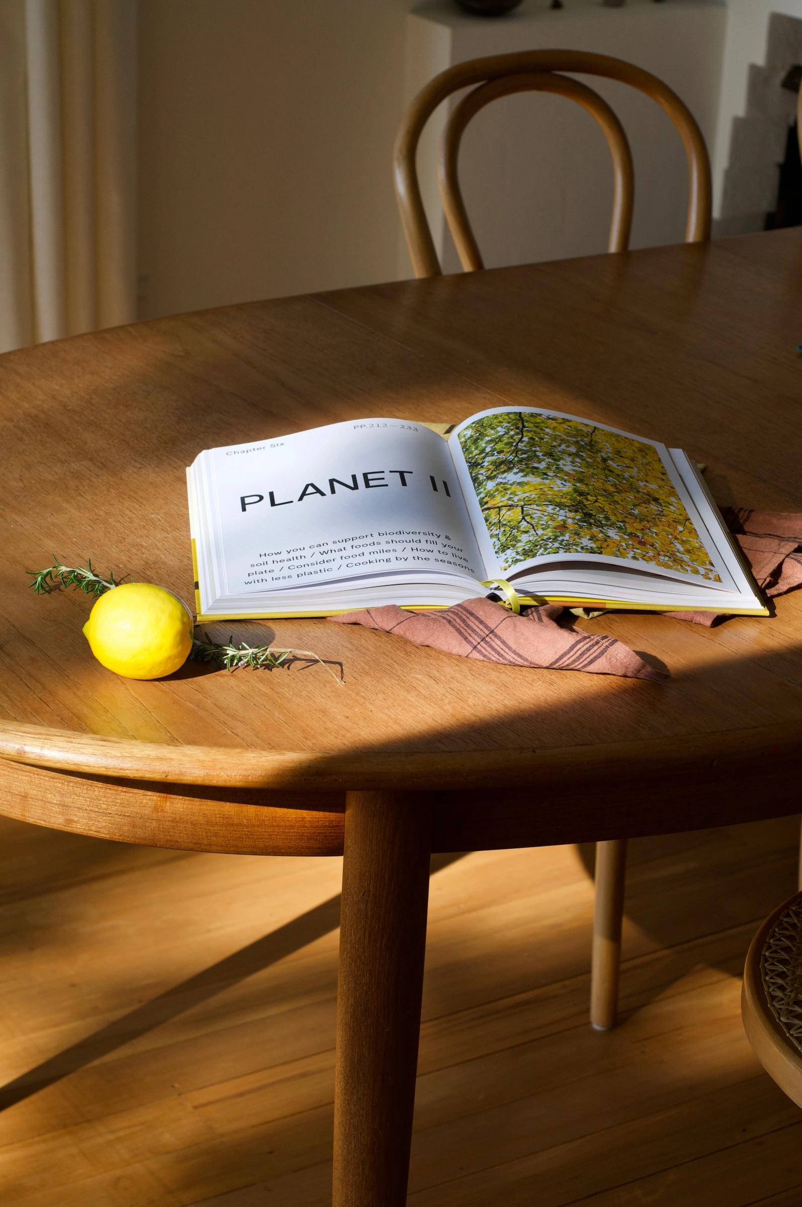 Open "PLANET II" book on wooden table, covering pink cloth. Lemon and greenery nearby. Chair in background.