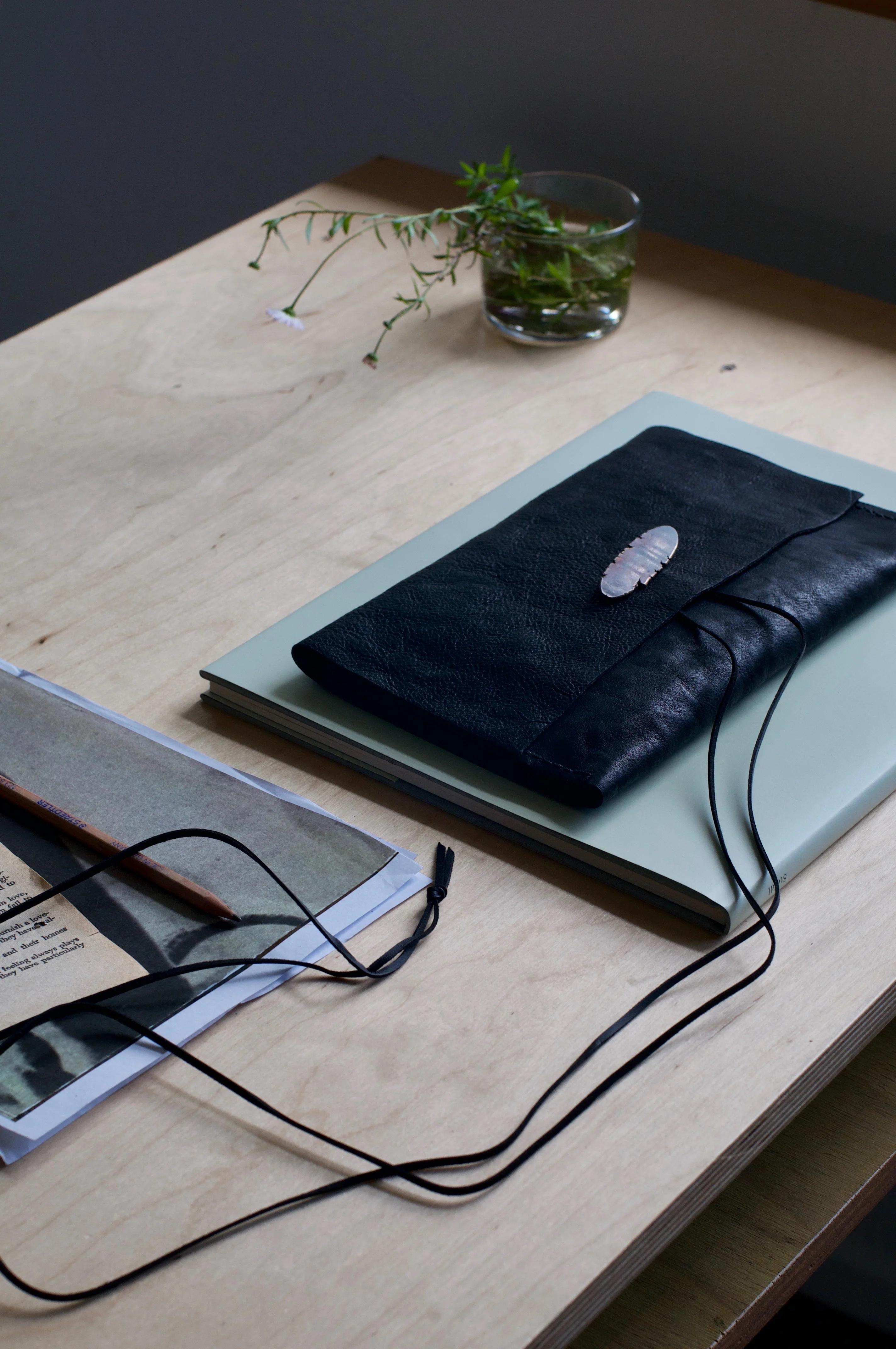 Black notebook on light green book. Open magazine with pencil nearby. Small glass with water and plant stems in background.