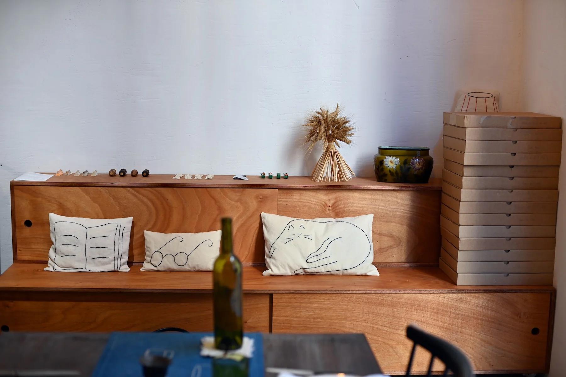 Wooden bench with cushions featuring minimalist designs. Pizza boxes, vase with wheat, marbles, and wine bottle on table.