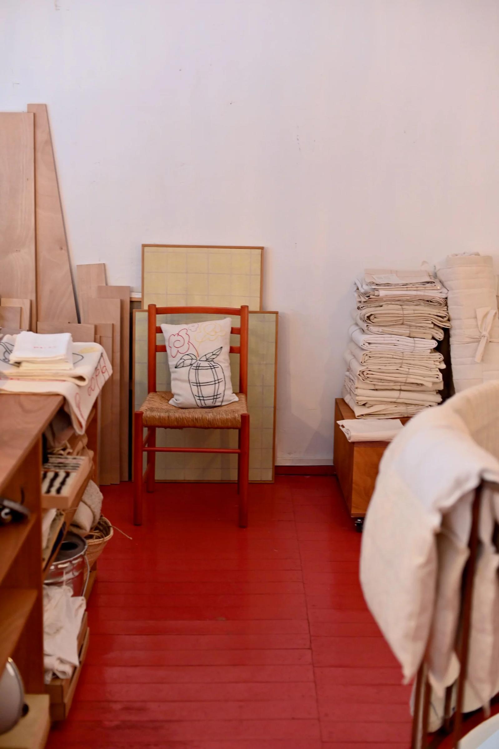 Room with red floor, wooden chair, and stacked fabrics. Shelves with various items on left.