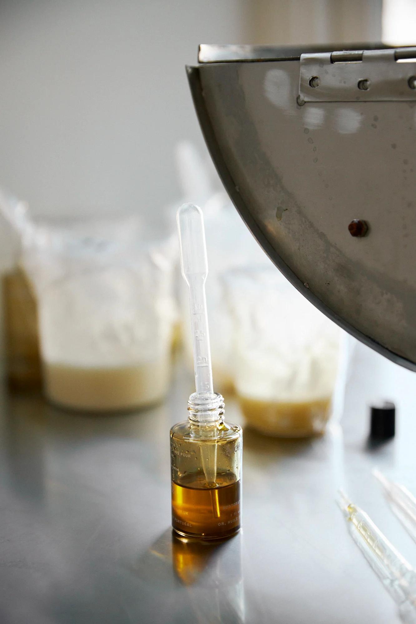 Small glass dropper bottle with amber liquid on metal surface. Blurred containers and metal object in background.