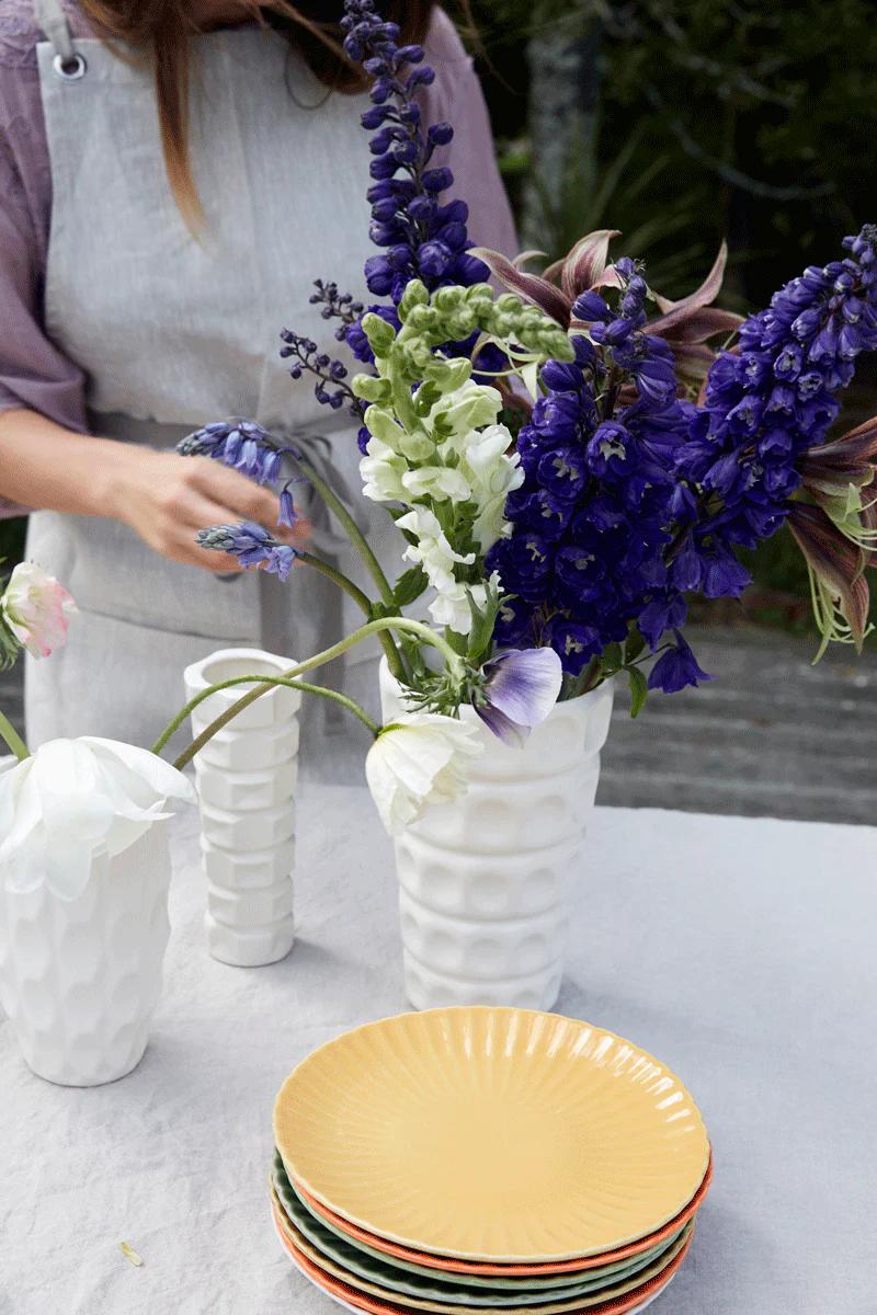 A person in a grey apron arranges purple and white flowers in a textured vase. Stacked colourful plates and smaller vases adorn the table, perfect for a Rachel Carley dinner party.