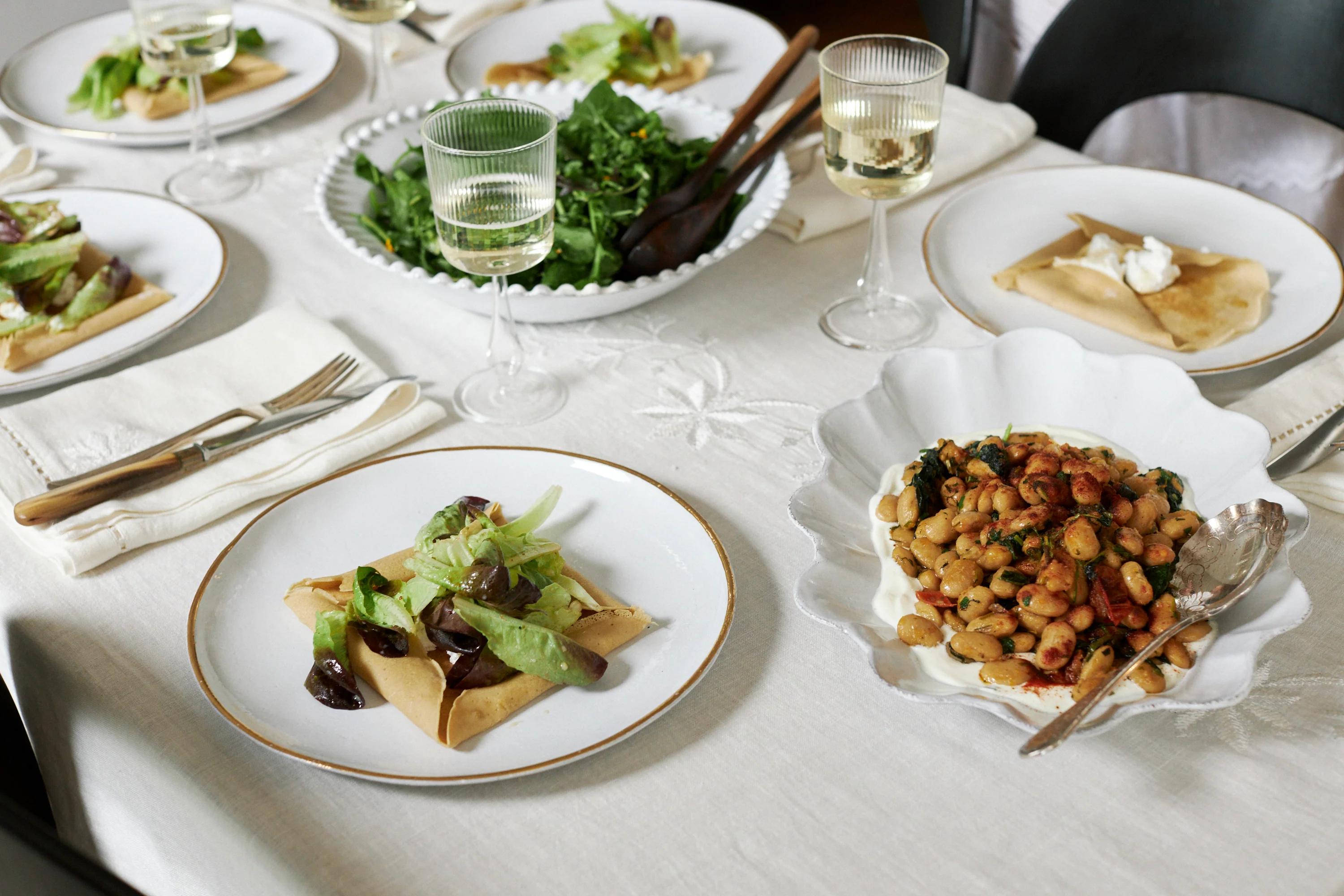 A table set for Easter with salads, flatbreads, beans, and wine. A large salad bowl centres the scene, completed with white linens and silverware.