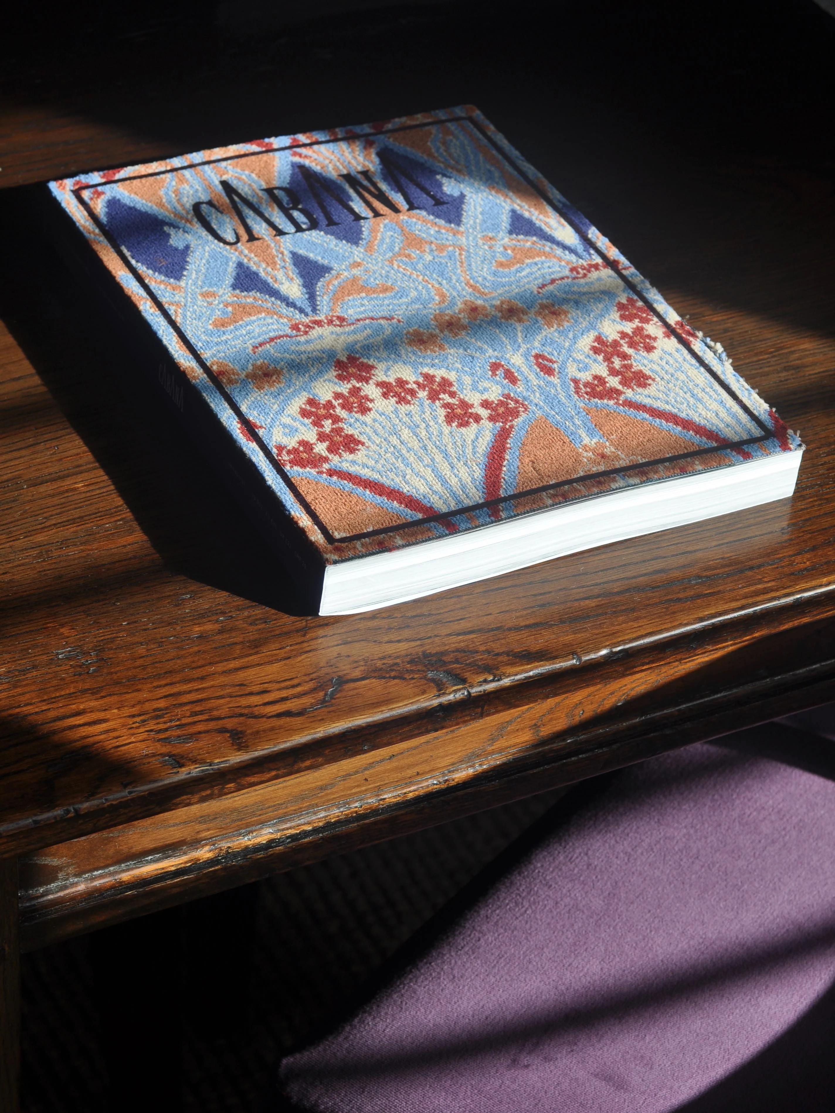 Book with ornate floral cover on wooden surface. Sunlight illuminating part of book. Purple cloth visible near bottom.