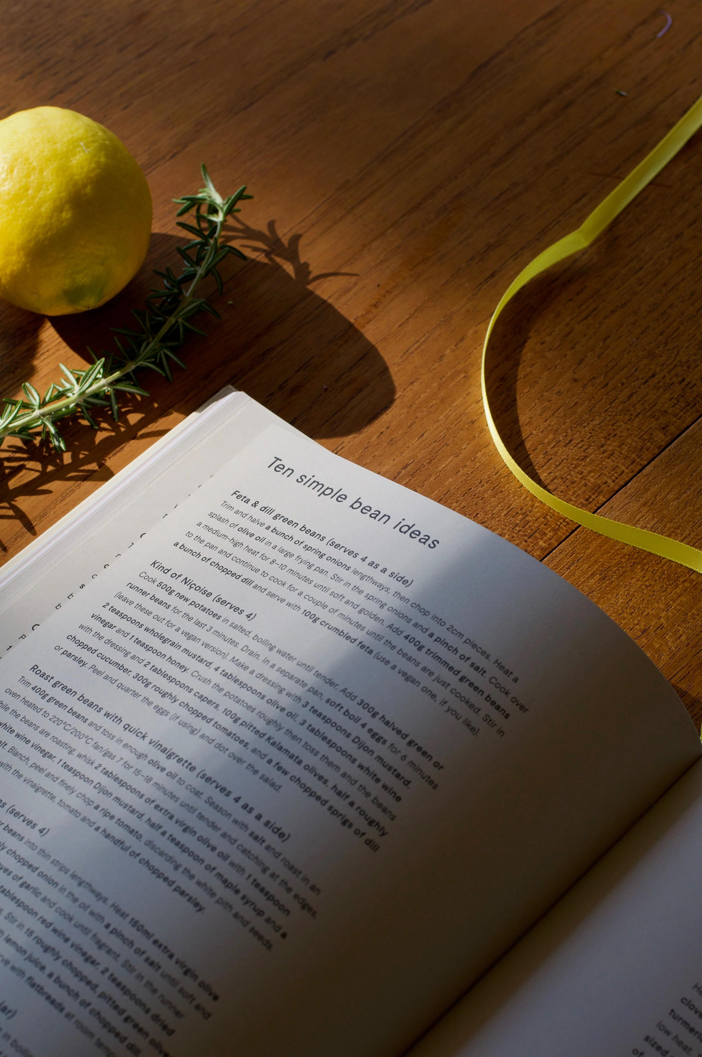 Open cookbook on wooden table. "Ten simple bean ideas" page visible. Lemon, rosemary, yellow ribbon nearby.