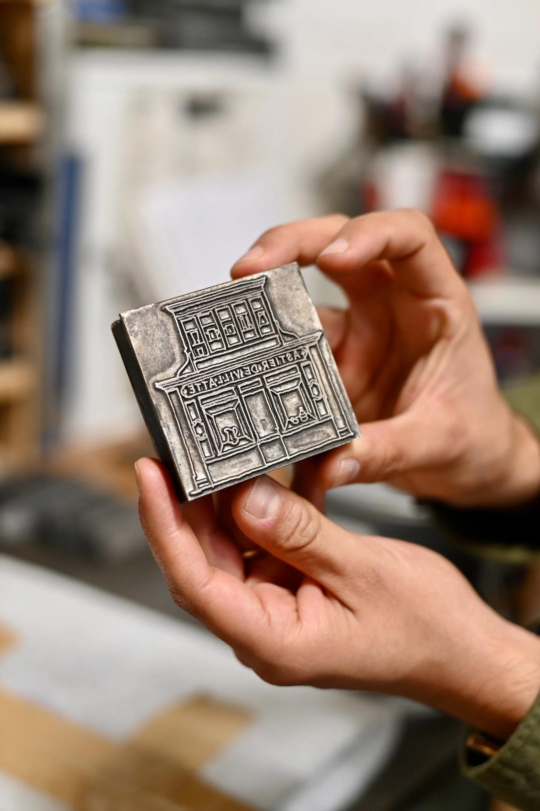 Two hands holding small, detailed metal embossing stamp with intricate storefront design.