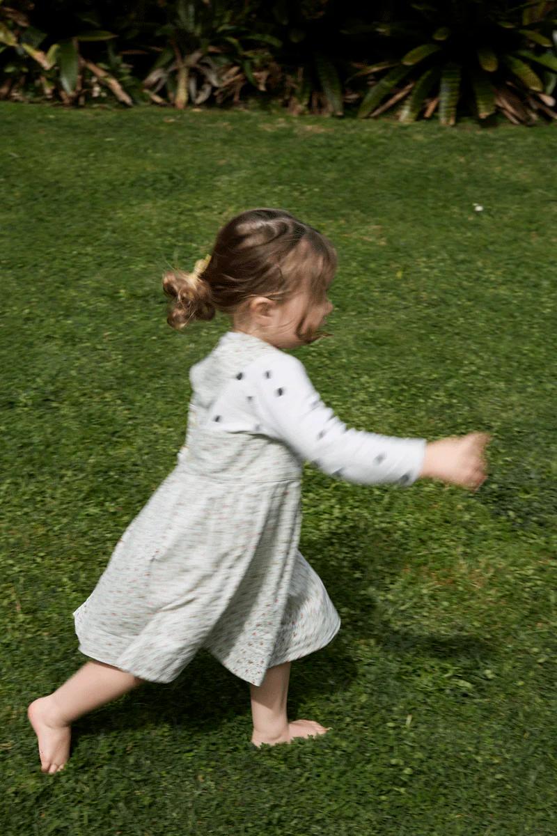 Child with hair in buns runs barefoot on grass. Wearing long-sleeve shirt and light dress. Captures joyful holiday moment.