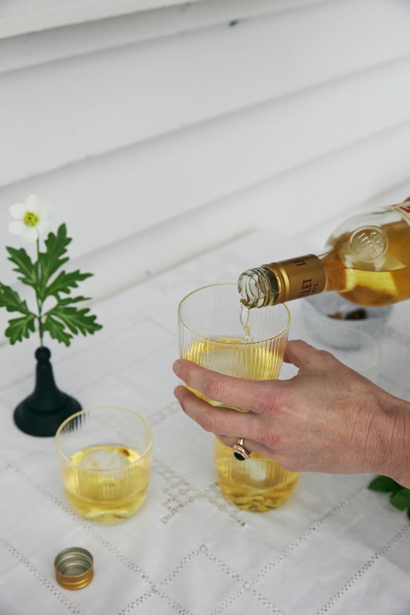 Hand pouring light drink into glass. Flower vase, another filled glass, bottle cap, and small bowl visible.