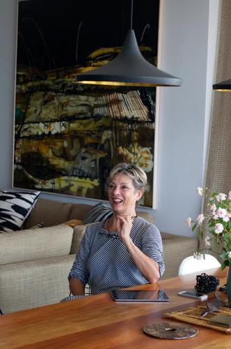 A person with short hair sits at a wooden table, smiling and gesturing. An abstract painting hands on the wall behind them, as well as a beige sofa with pilloe, and a vase of flowers on the table. Modern pendant lights hang from the ceiling.