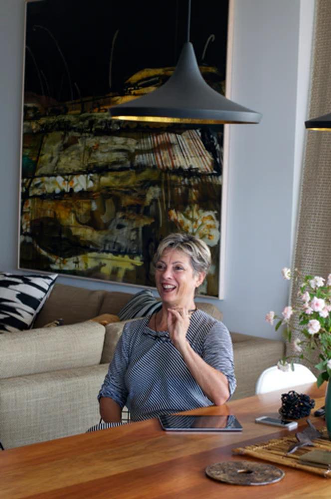 A person with short hair sits at a wooden table, smiling and gesturing. An abstract painting hands on the wall behind them, as well as a beige sofa with pilloe, and a vase of flowers on the table. Modern pendant lights hang from the ceiling.