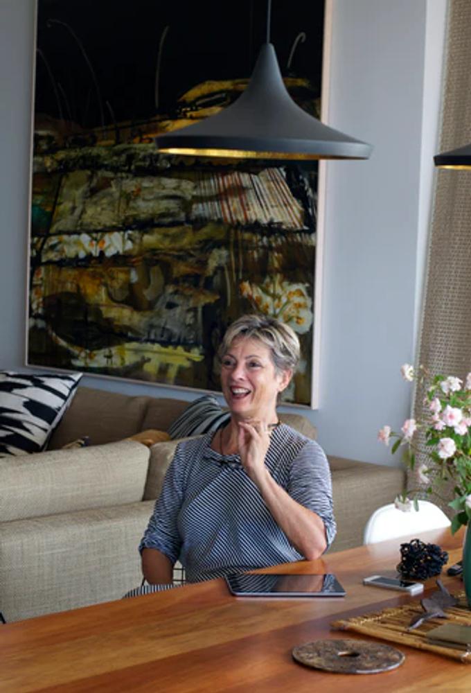 A person with short hair sits at a wooden table, smiling and gesturing. An abstract painting hands on the wall behind them, as well as a beige sofa with pilloe, and a vase of flowers on the table. Modern pendant lights hang from the ceiling.