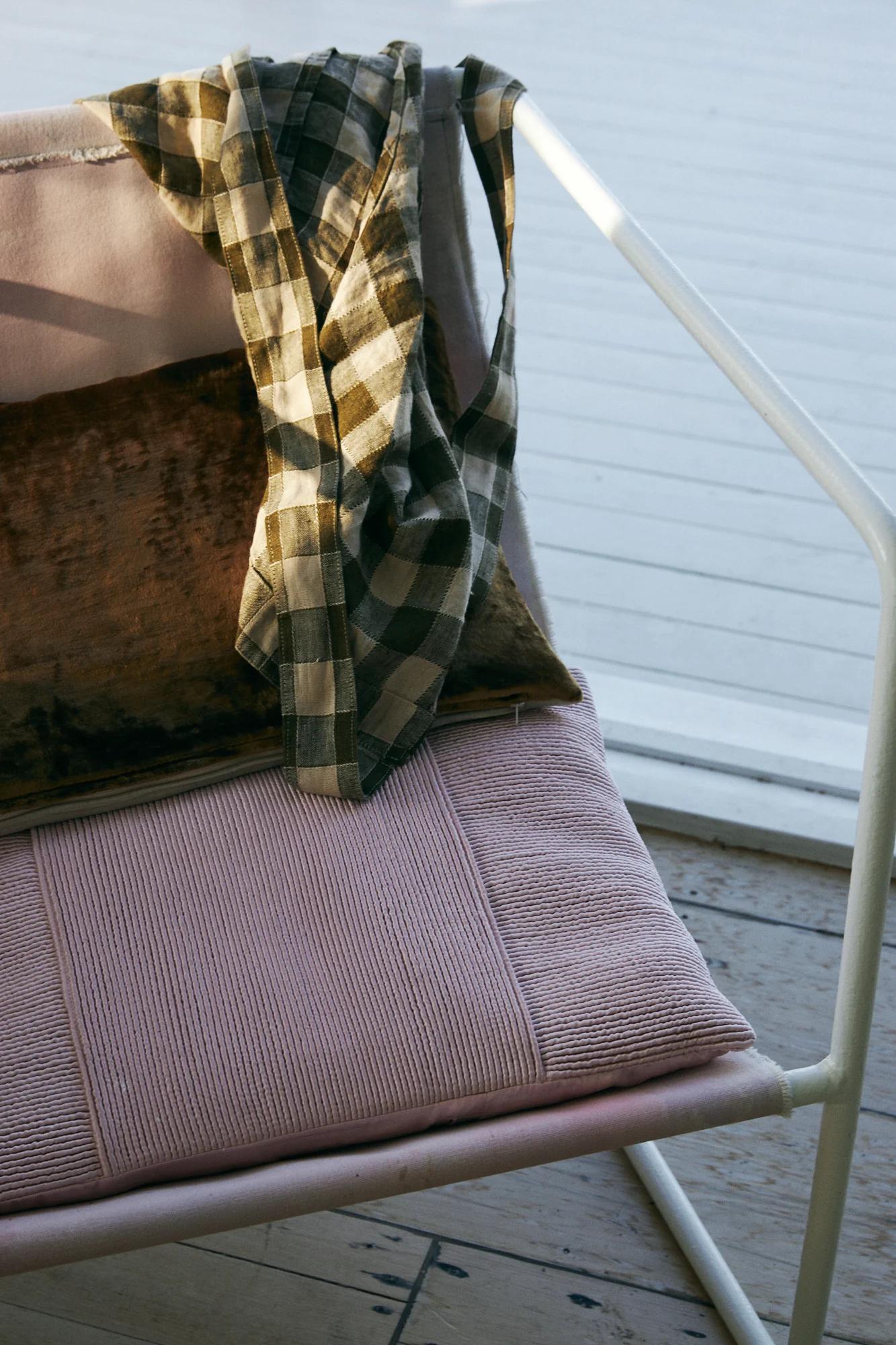 Pink cushioned chair by window. Brown checkered shirt draped over. Brown pillow on seat. Sunlit scene.