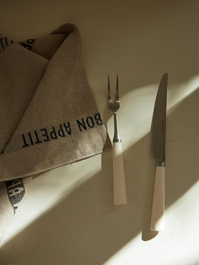 Knife and fork with white handles on light table. Folded "BON APPÉTIT" napkin nearby. Sunlit scene.